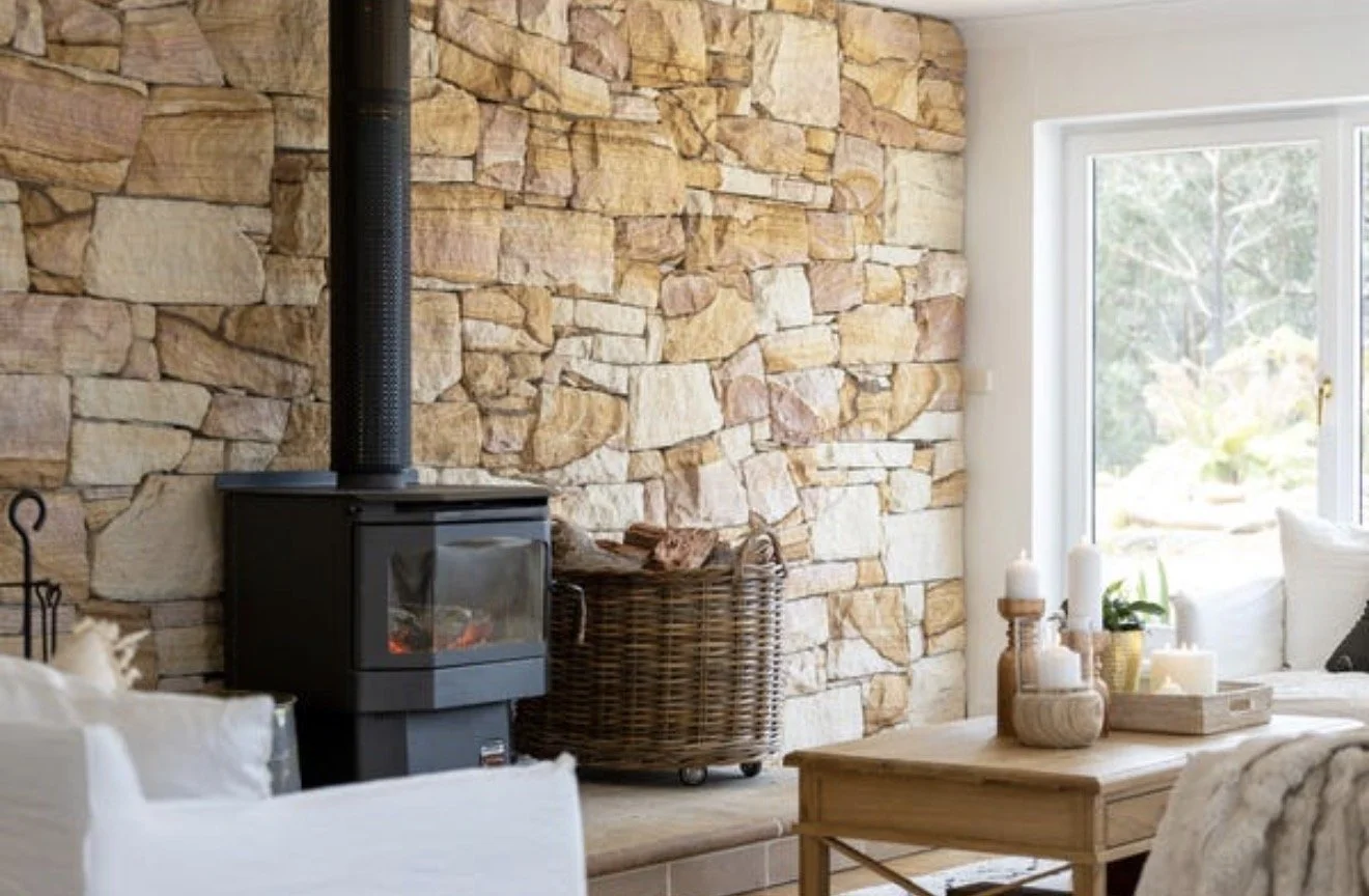 A cozy living room with a stone accent wall, a black wood-burning stove, wicker basket, wooden coffee table with candles, and a large window with a view of greenery outside.