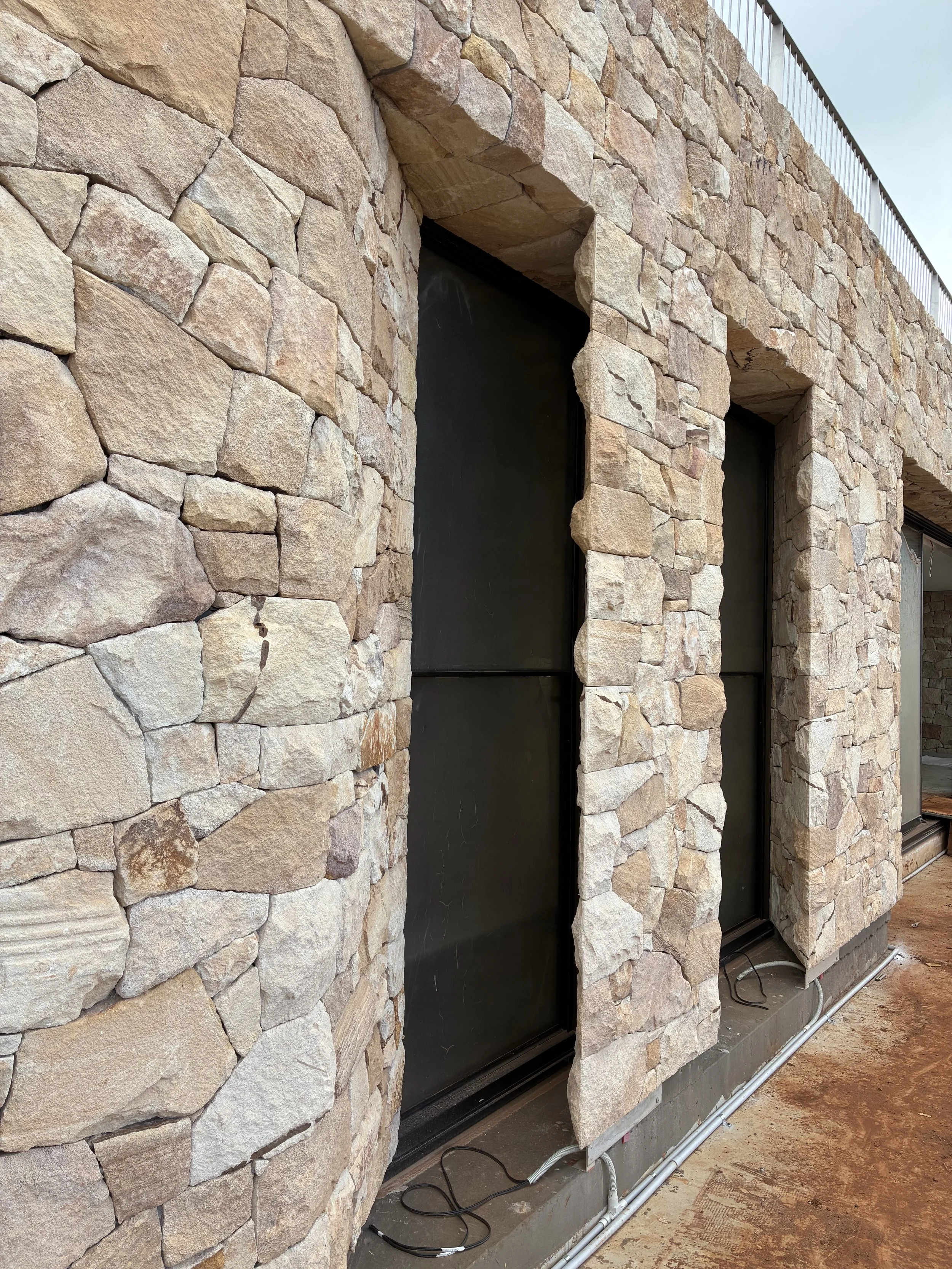 Close-up of a stone wall with cutouts for windows or doors, showing black panels, electrical wiring, and plumbing at the base, under a construction or renovation site.