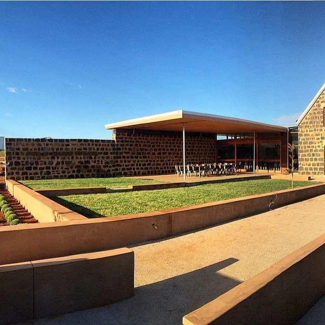 Modern building with a slanted roof, stone walls, large windows, and a patio with chairs, surrounded by a landscaped garden with green grass and wooden borders under a clear blue sky.