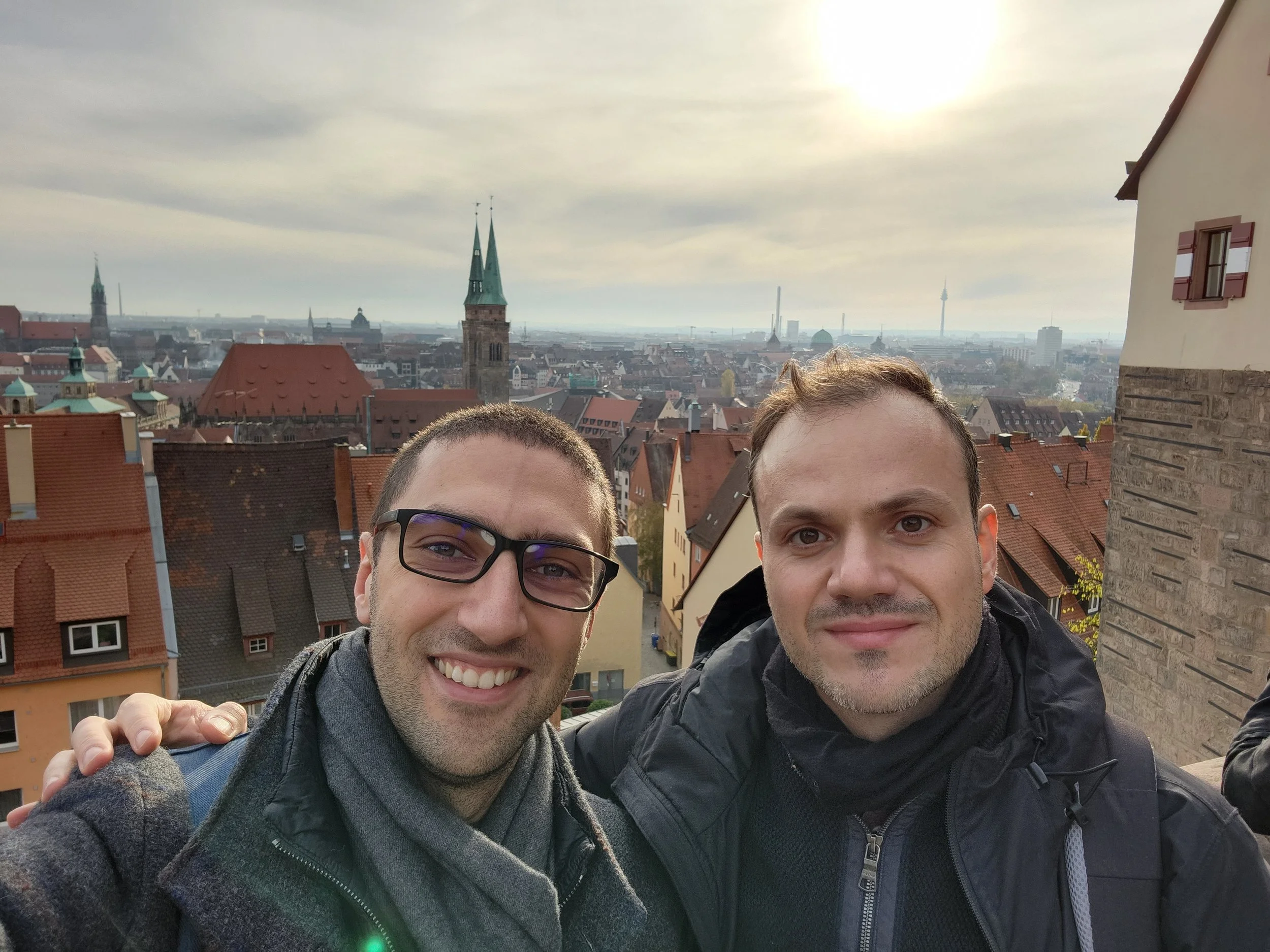 Two smiling men taking a selfie with a cityscape in the background, including rooftops, church steeples, and a tall tower, on a cloudy day.