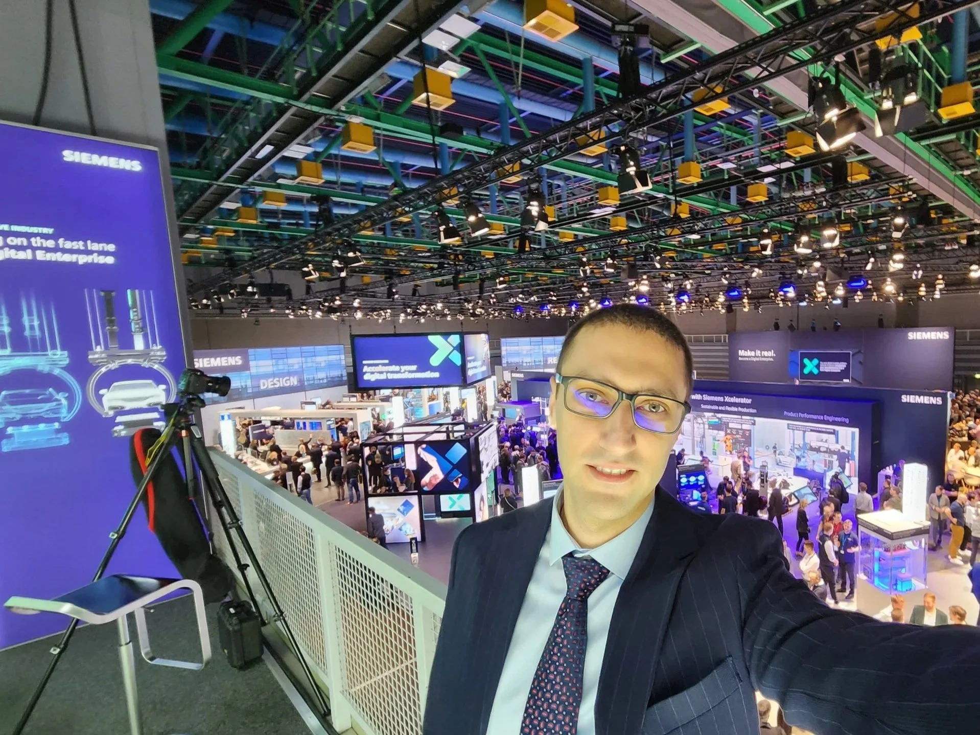 A man wearing glasses, a dark pinstripe suit, and a tie taking a selfie at a SPS Messe Nuremberg exhibition. The background features numerous displays, screens, and a crowd of people, with lighting and booth setups indicating a professional event.