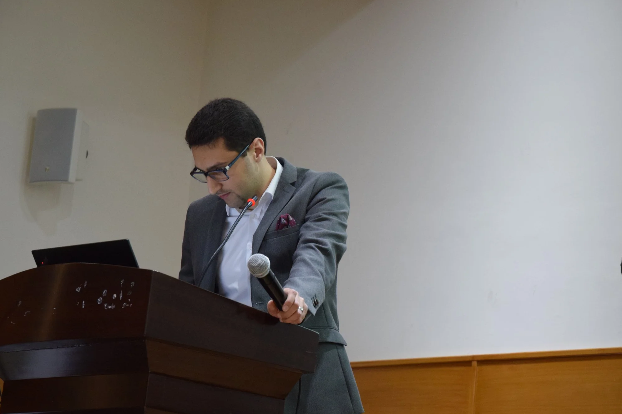 Rami Abuodeh with dark hair and glasses wearing a gray suit and white shirt is standing behind a wooden podium, looking down at a laptop, holding a microphone in his right hand. There is a white wall with a speaker mounted on it behind him.