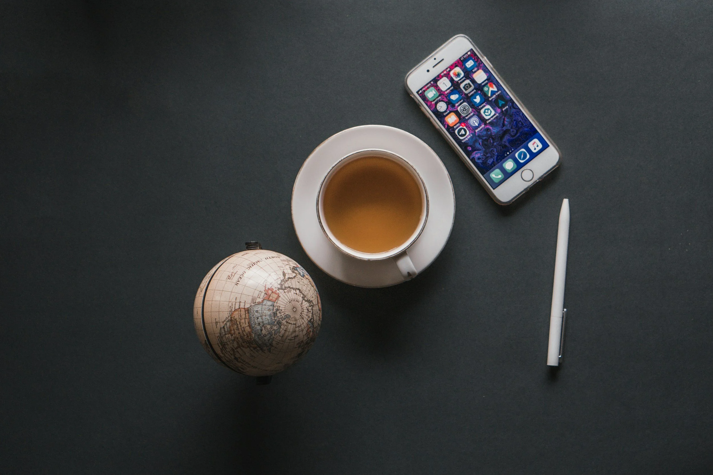 A dark surface with a globe, a white coffee cup filled with tea on a saucer, an iPhone with colorful apps on the screen, a white stylus, and a small globe of Earth.