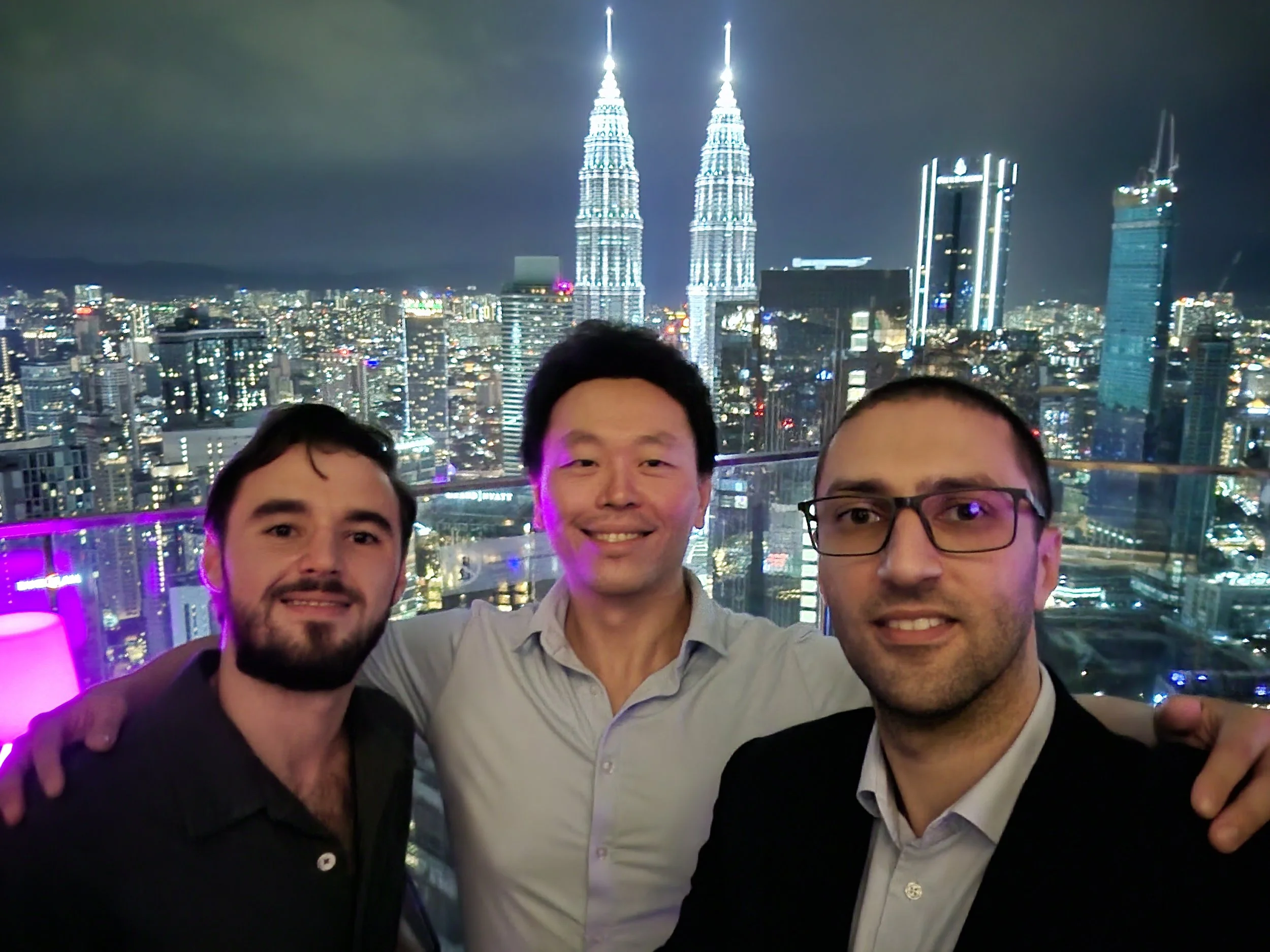 Three men smiling for a selfie at night with the Kuala Lumpur skyline and the Petronas Twin Towers illuminated in the background.