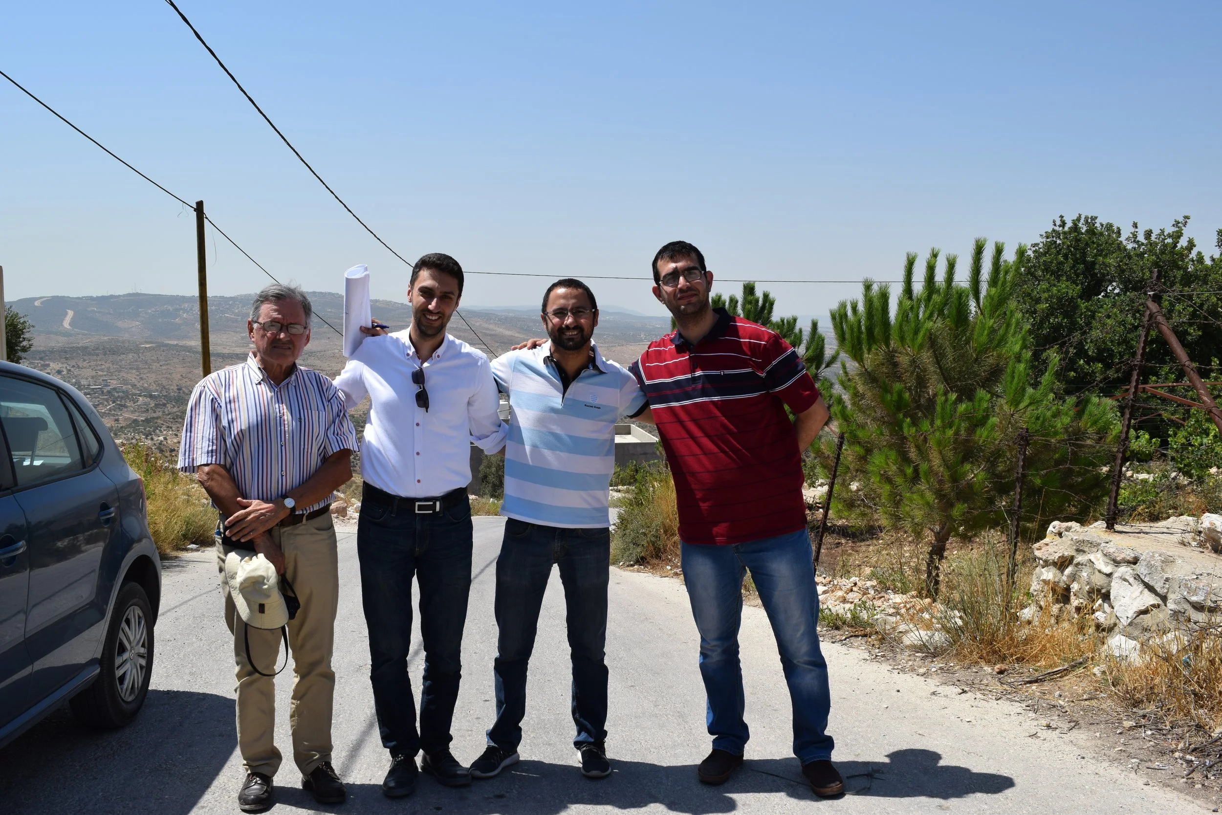 Four men standing outdoors on a sunny day, smiling at the camera. They are dressed casually, with hills and sparse vegetation in the background. One man is holding a rolled-up document or paper. Yamoun Project.