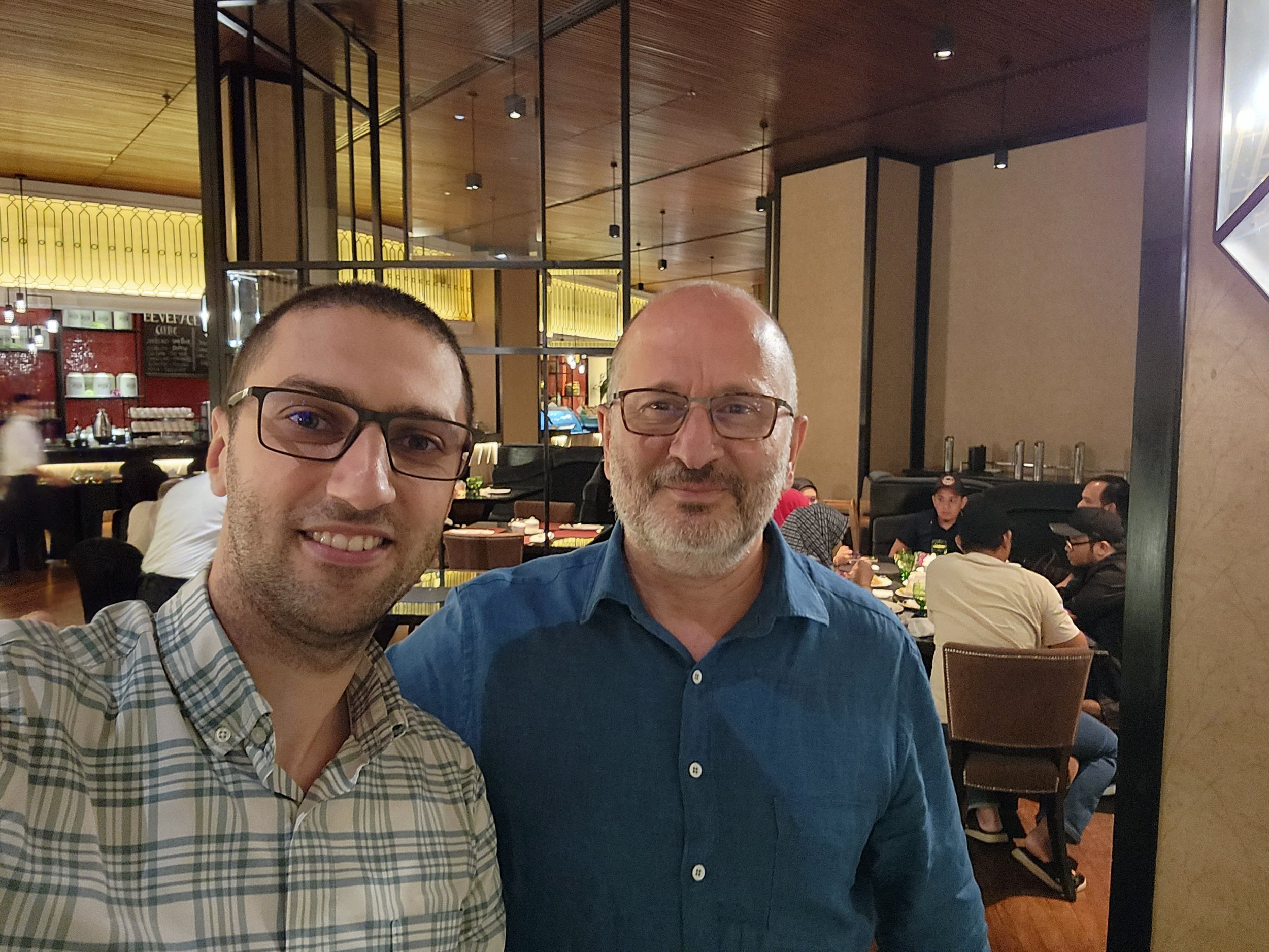 Two men smiling and taking a selfie inside a restaurant. The man on the left has short dark hair, wears glasses and a checkered shirt. The man on the right has a beard, wears glasses and a blue shirt. The restaurant has warm lighting, wooden accents,