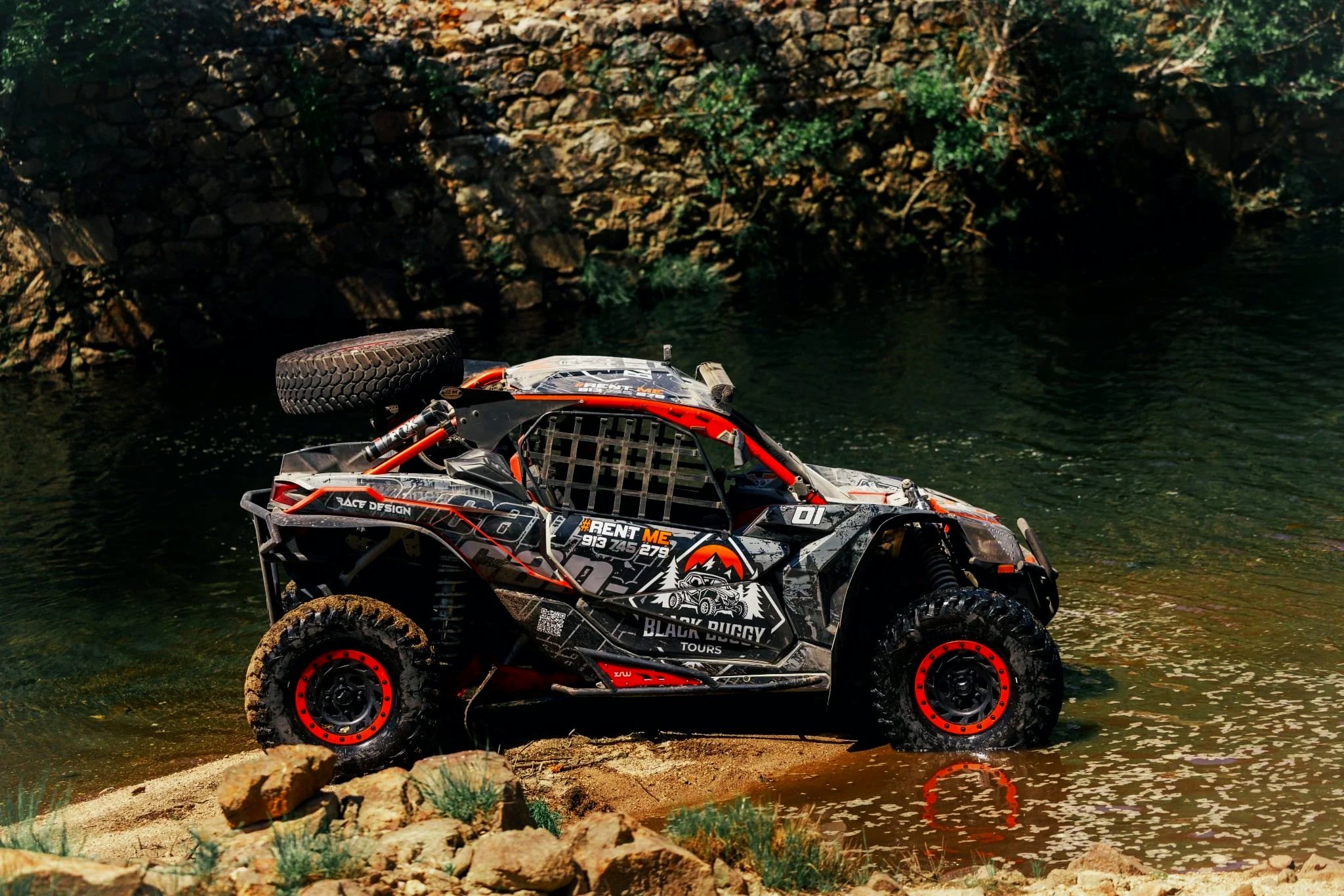 Veículo off-road com rodas grandes e pneus sujos, parcialmente submerso na água de um rio, com paredes rochosas ao fundo e vegetação ao redor.