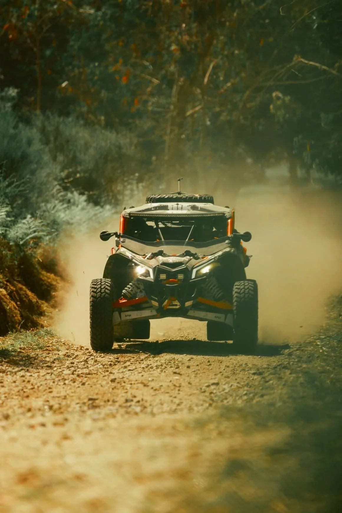 Veículo off-road branco e preto acelerando por uma estrada de terra, com poeira levantando atrás e árvores ao redor, durante o dia.