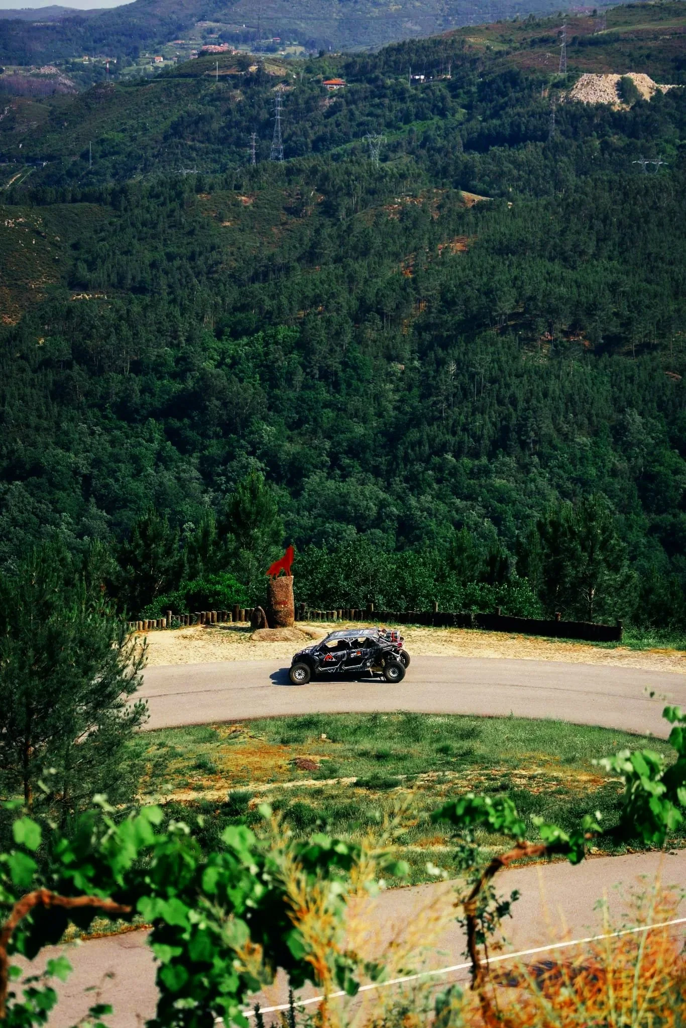 Carro preto estacionado ao lado de uma cerca de arame em uma área com muitas árvores e vegetação, com colinas verdes ao fundo.