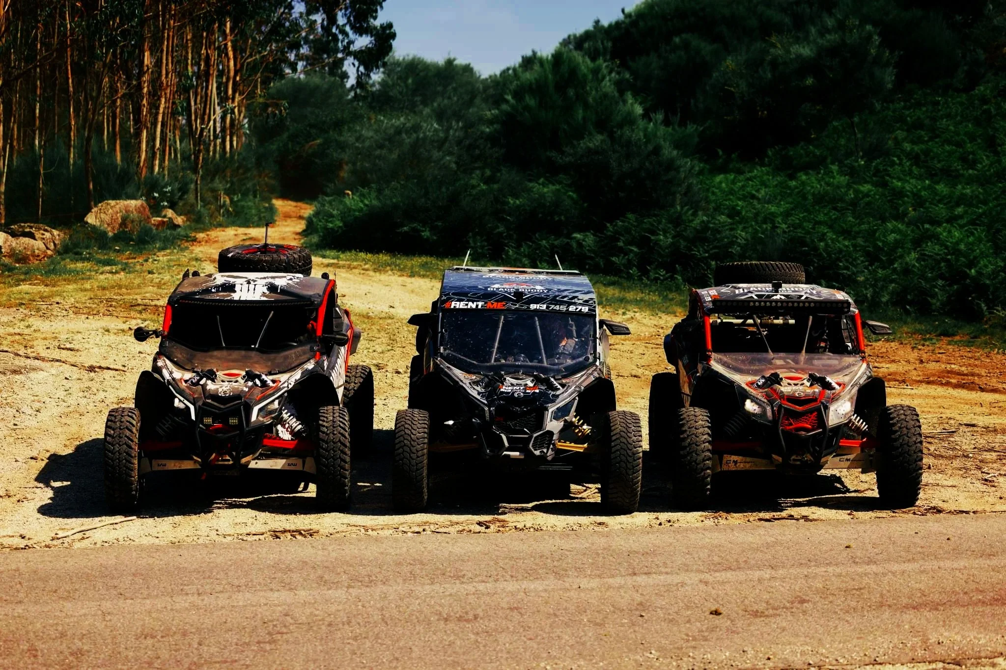 Três veículos off-road em linha em uma estrada de terra, rodeados por vegetação de árvores e arbustos.