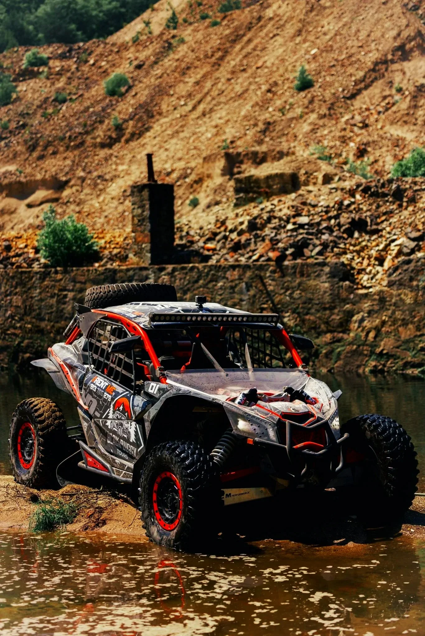 Veículo de rally off-road preto, branco e vermelho atravessando uma água em um ambiente de terreno rochoso e árido.