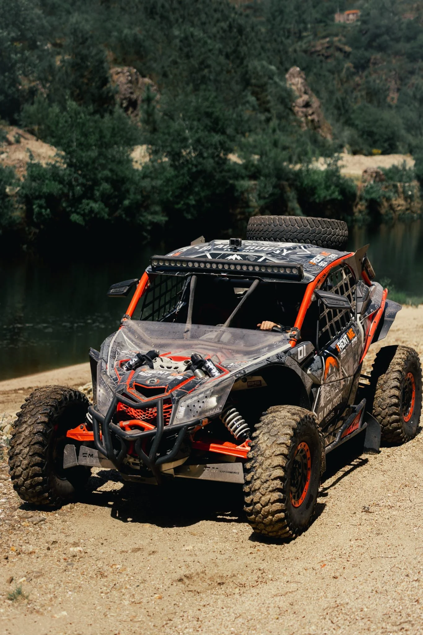 Veículo off-road com rodas grandes, pneus sujos e estrutura de proteção, estacionado em uma área arenosa próxima a um rio ou lago, com vegetação e colinas ao fundo.