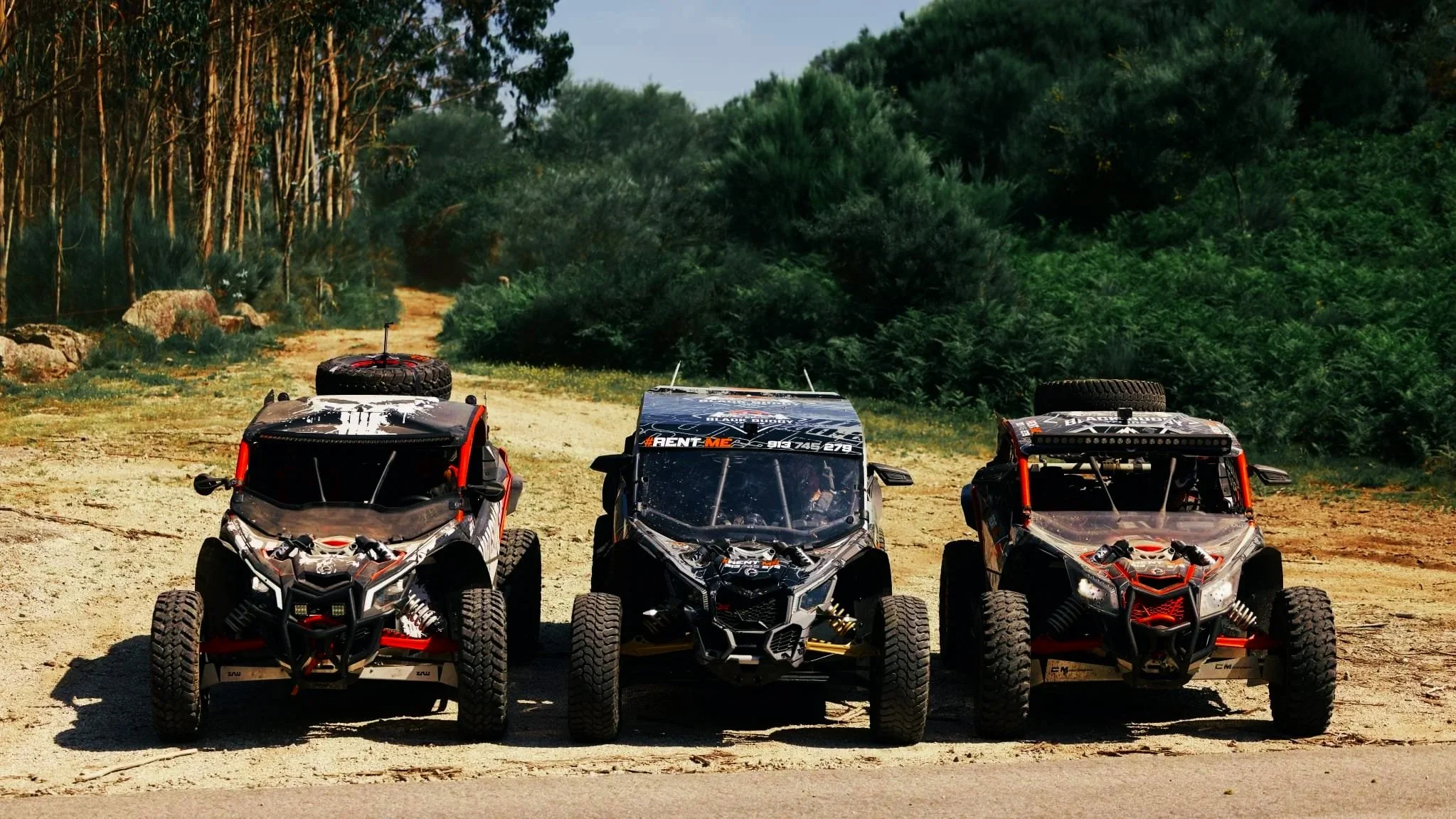 Três veículos off-road em uma trilha de terra, rodeados por vegetação verde, com árvores ao fundo.