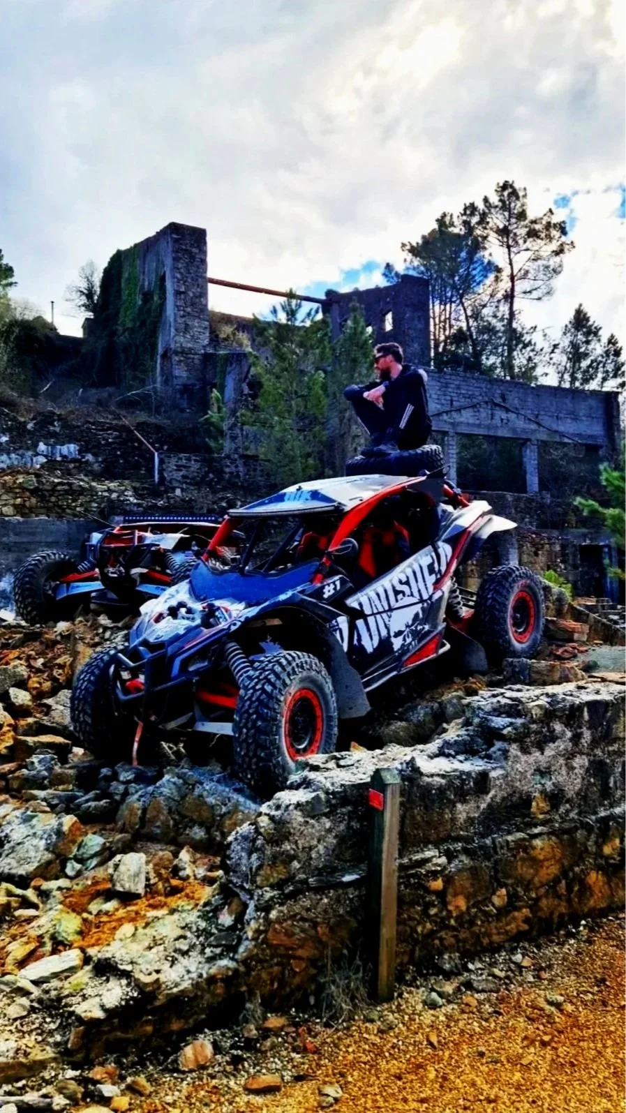 Um carro de corrida off-road em um terreno rochoso, com uma pessoa sentada no topo da estrutura do carro e uma paisagem de árvores e ruínas ao fundo.