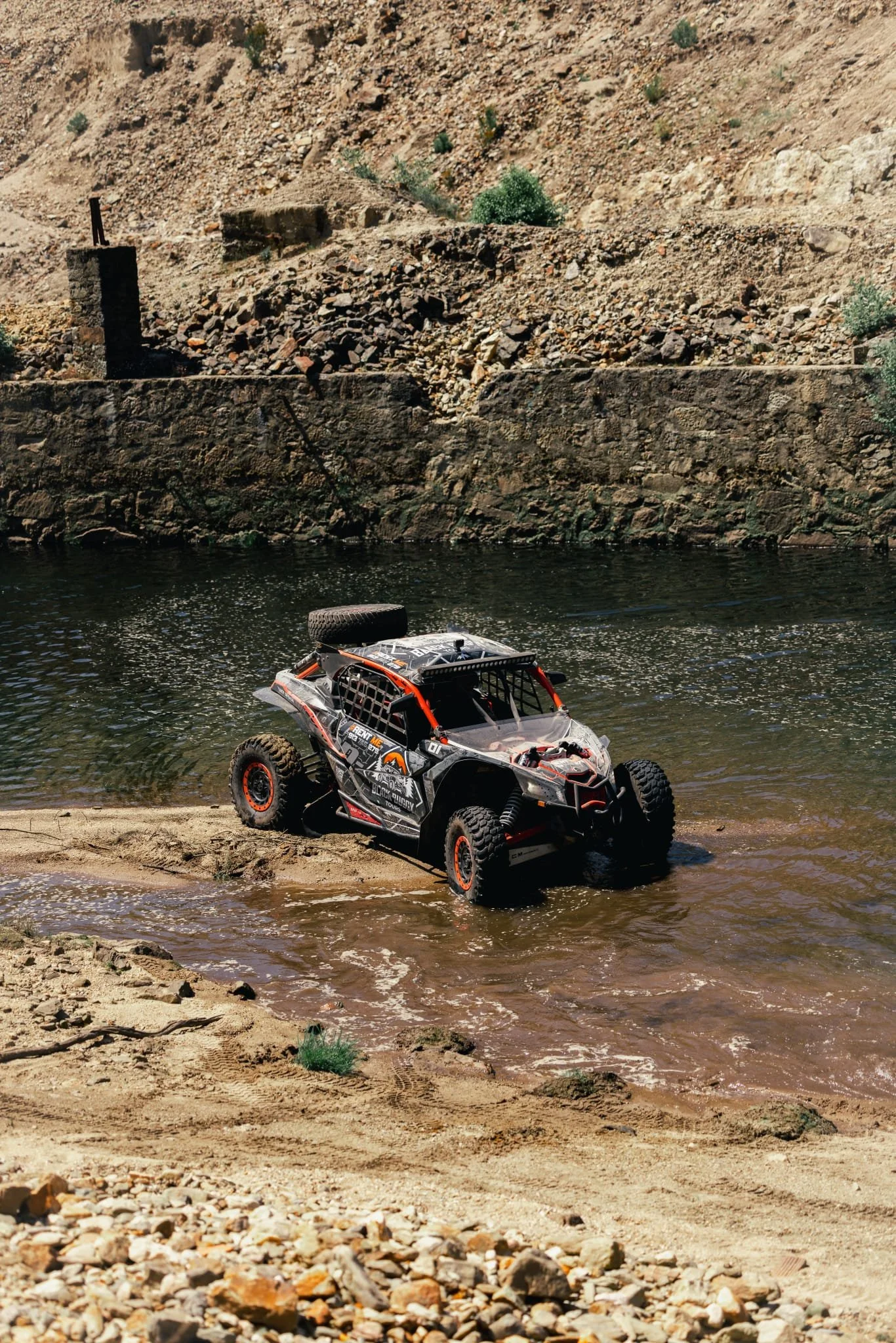 Veículo off-road atravessando um rio em um ambiente desértico, com muitas pedras e vegetação escassa ao redor.