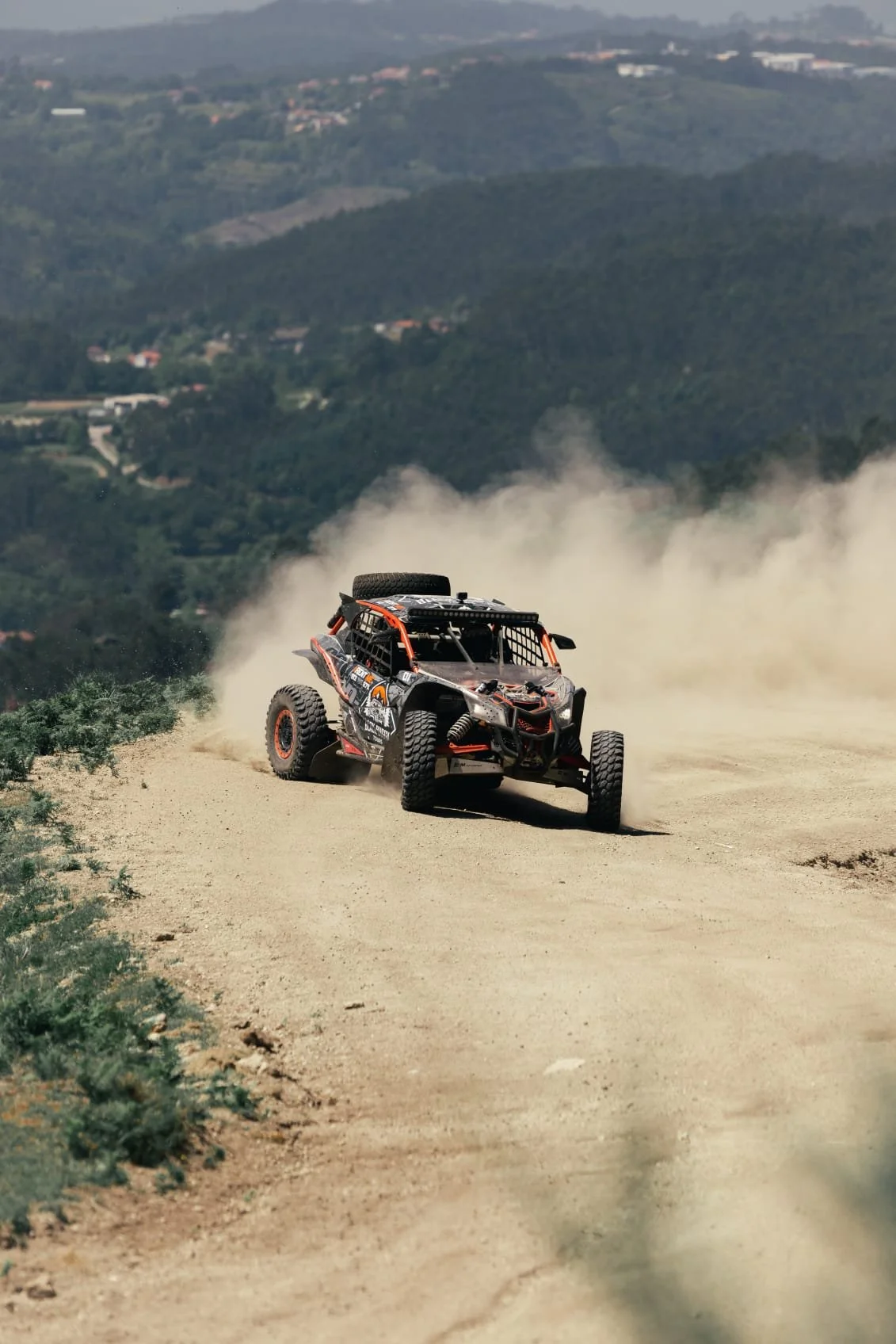Veículo de corrida off-road segundo plano, dirigindo por um caminho de terra e levantando poeira, com uma paisagem de colinas verdes e um céu claro ao fundo.
