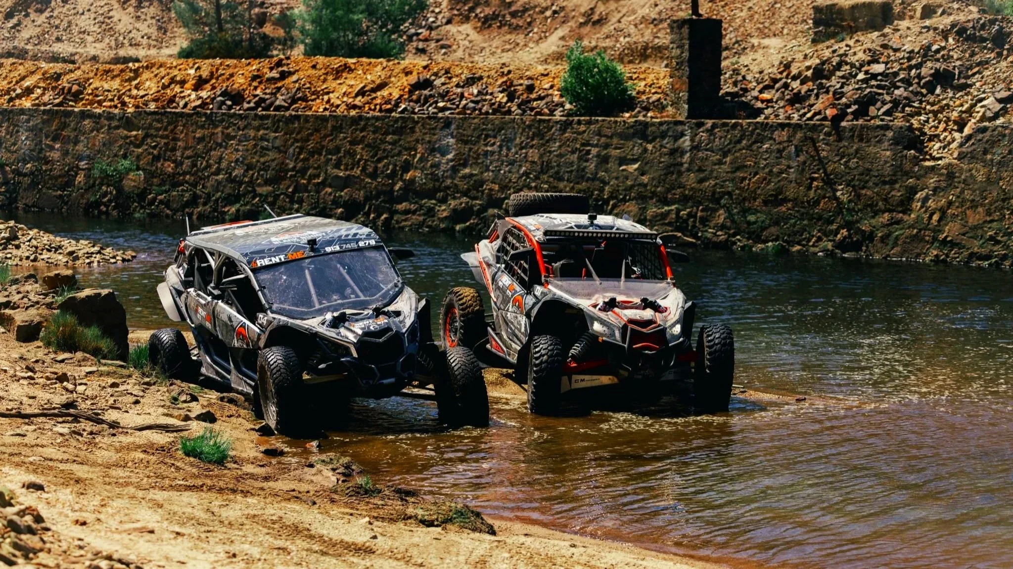 Duas buggys off-road coloridas próximas a um rio em um ambiente desértico com morros e vegetação escassa.