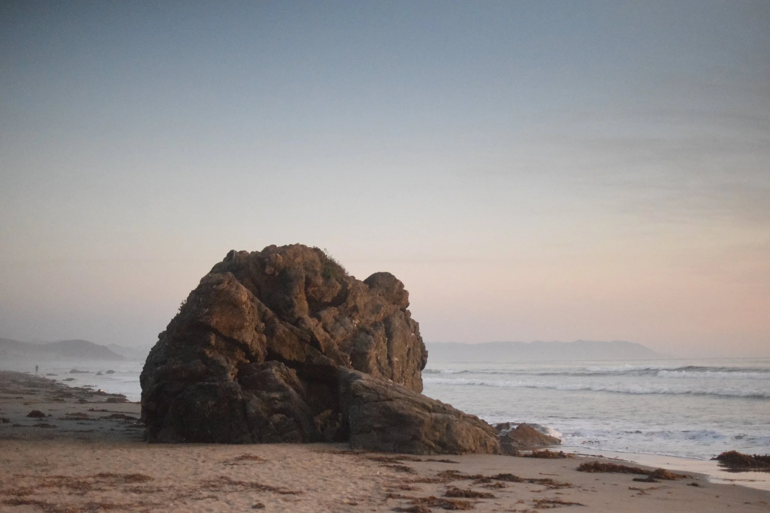 Large rock formation on a beach with the ocean in the background and a sky with soft shades of blue and pink during sunset or sunrise.