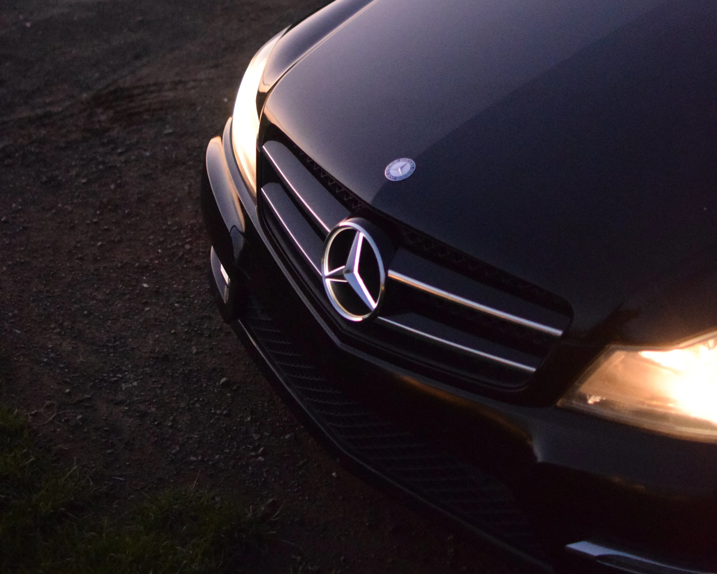 Close-up of the front of a black Mercedes-Benz car parked on a dirt surface, with its headlight and grille visible.