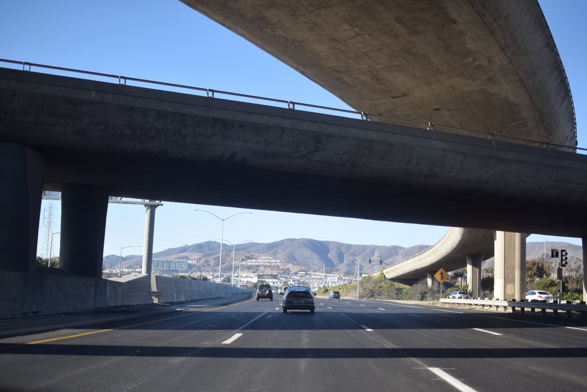 Multiple cars driving under an overpass on a highway with mountains in the background.