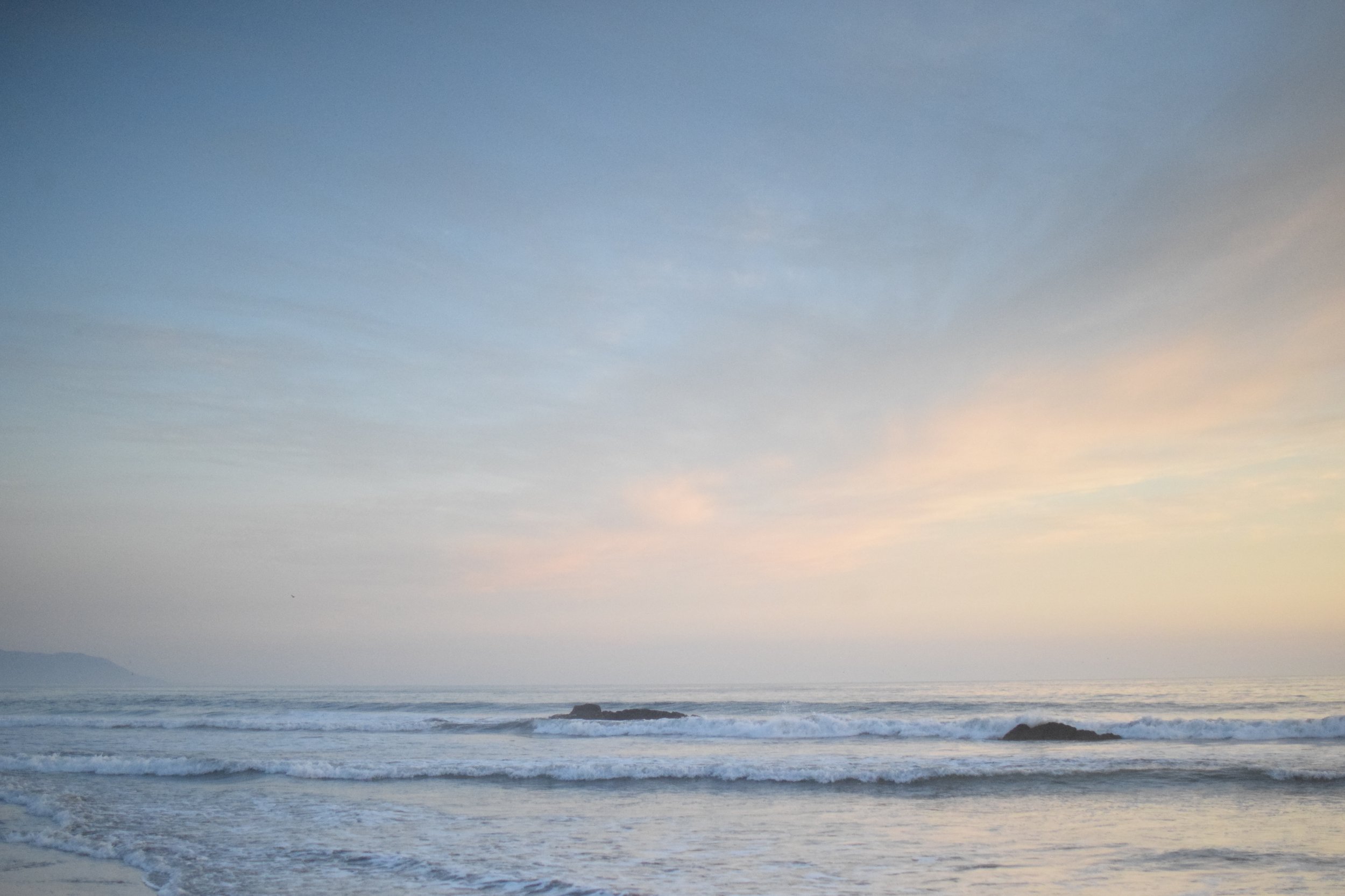 A beach scene at sunset with gentle waves, a calm sea, and a sky transitioning from blue to soft pastel colors near sunset.