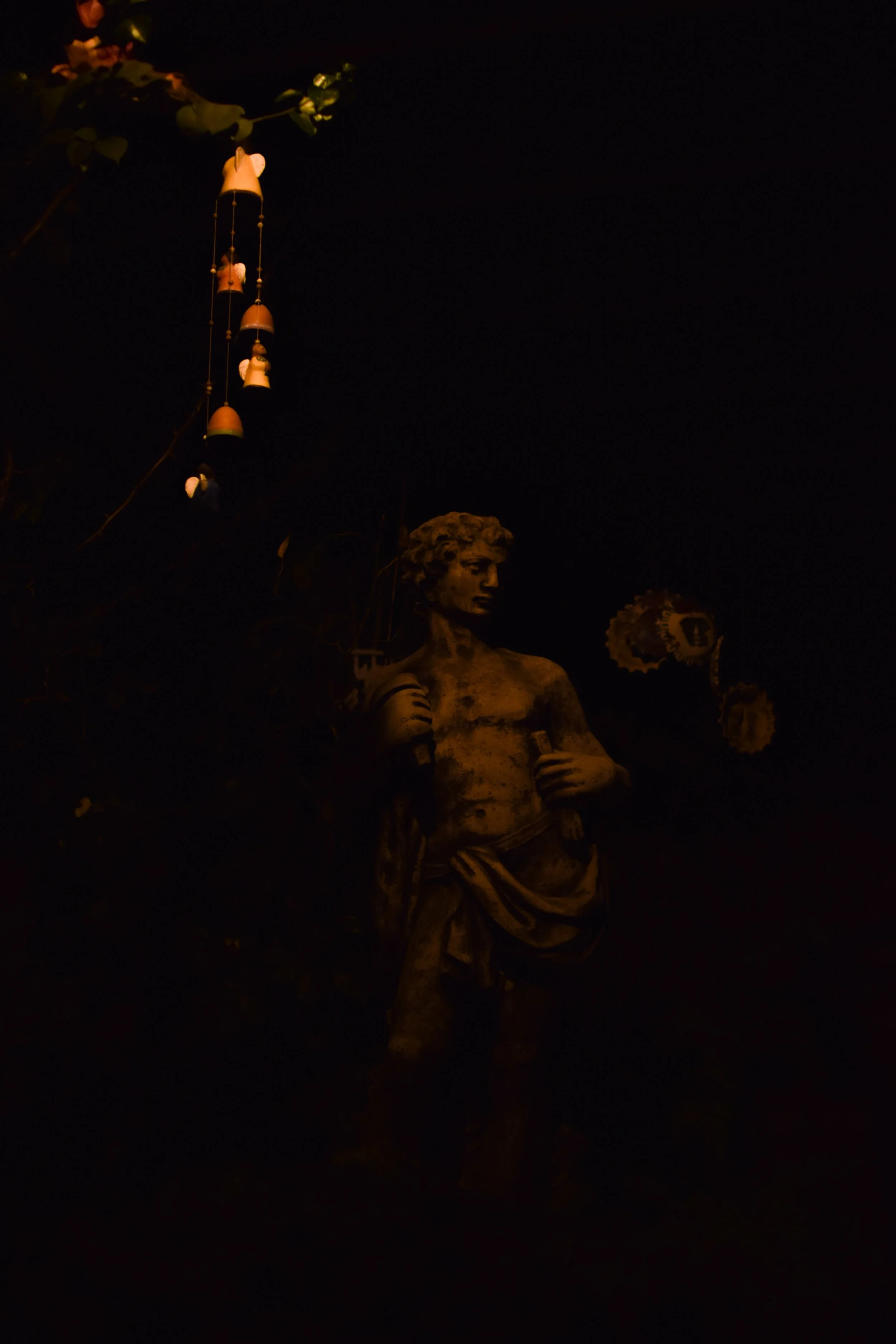 A statue of a person, likely a historical figure, holding a long object, illuminated against a dark background. There are decorative elements and possibly lanterns or hanging ornaments near the statue.