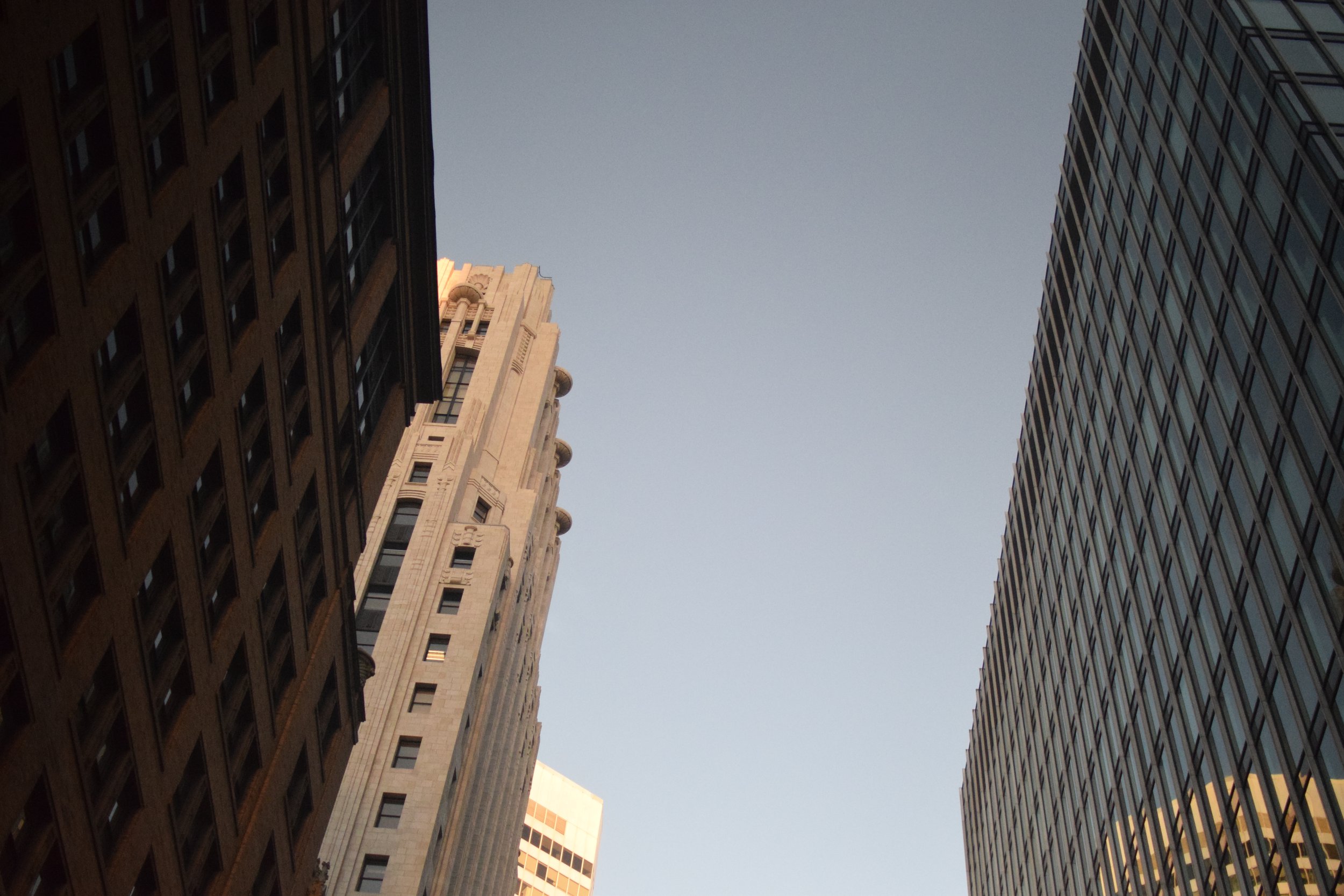 Photo taken from street level looking up at tall buildings in a city, showing a clear sky between the skyscrapers.