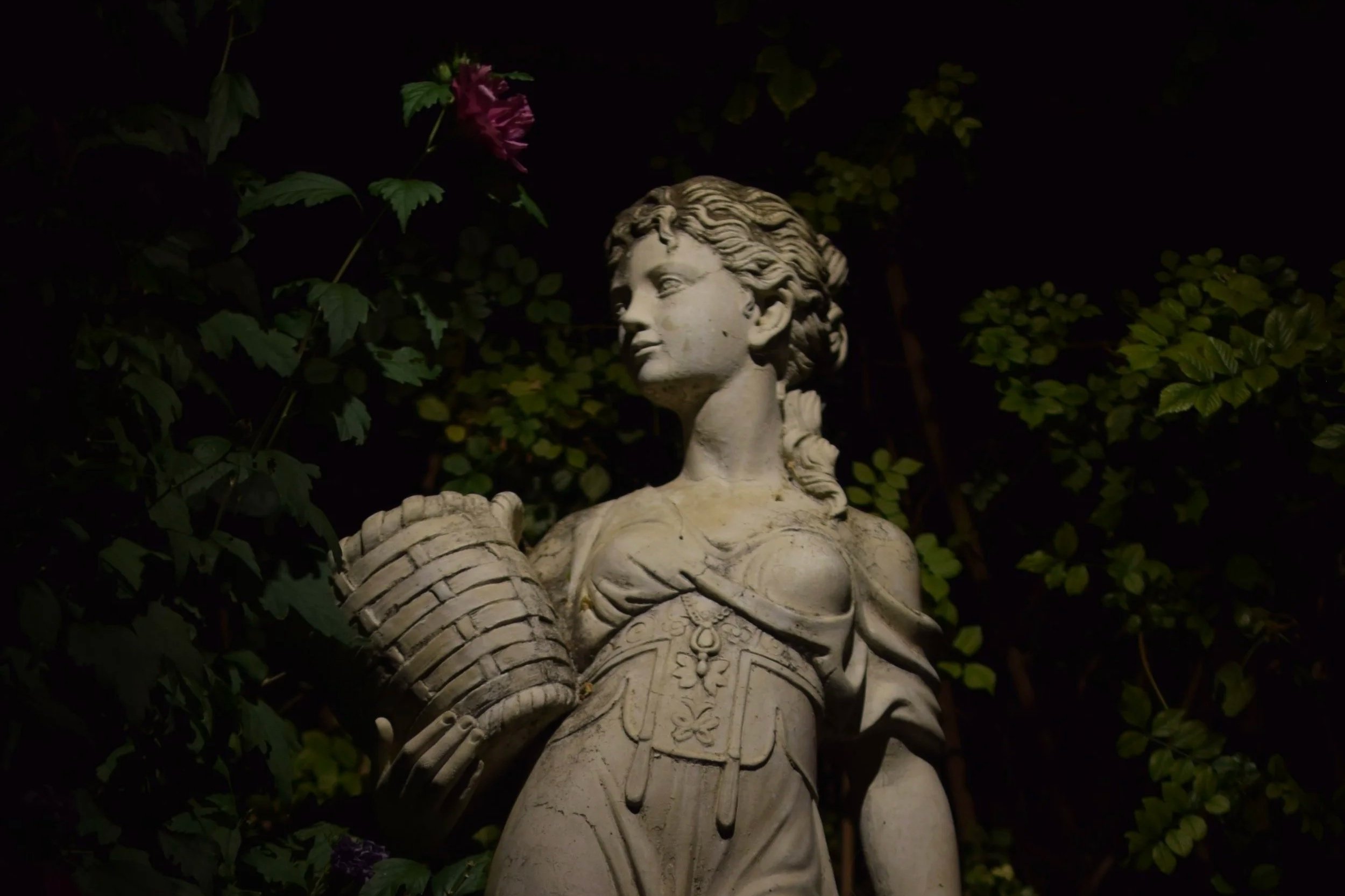 Nighttime photo of a stone statue of a woman dressed in historical clothing, surrounded by dark greenery and flowers.