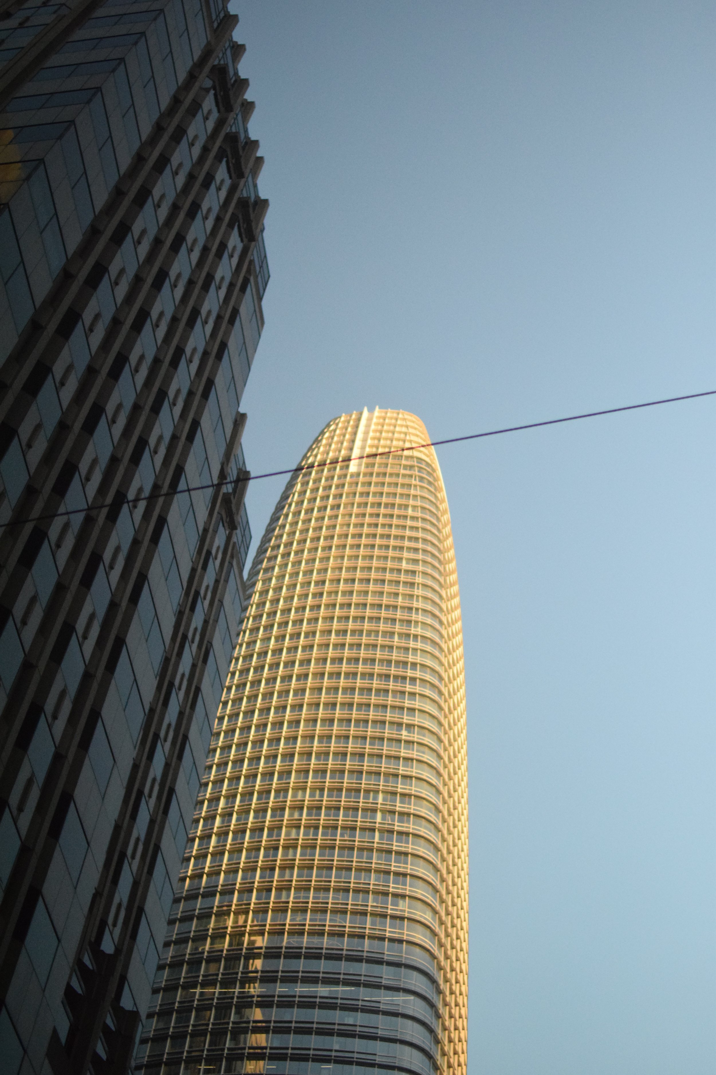Low angle view of two tall skyscrapers against a clear blue sky, one with reflective glass windows and the other with a rounded, grid-patterned facade, illuminated by warm sunlight.