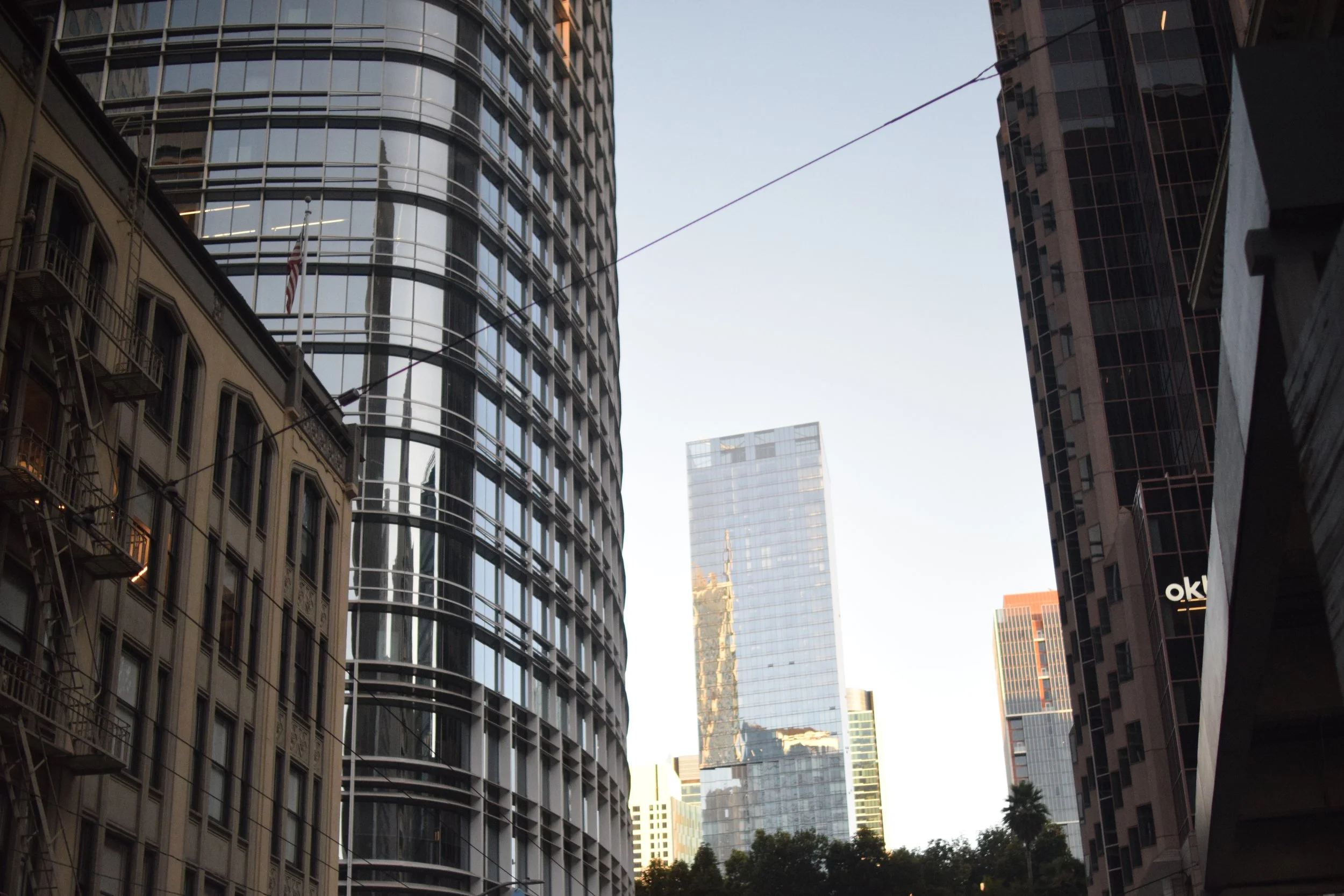 City street scene with skyscrapers, with glass buildings reflecting the sky and surrounding structures. A utility wire runs across the sky, and some greenery is visible at street level.