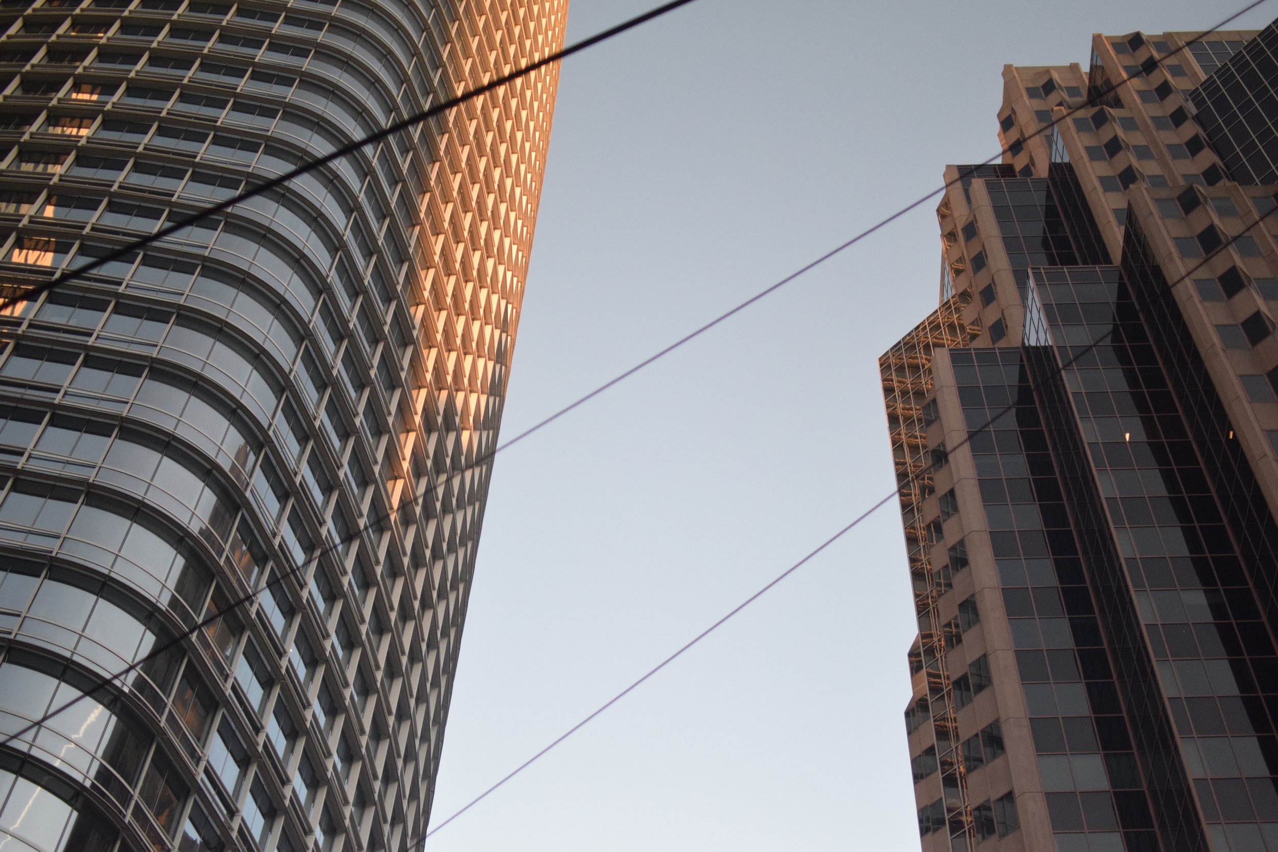 View of two high-rise buildings with glass and concrete exteriors, seen from below against a clear sky, with suspension cables crossing in the foreground.