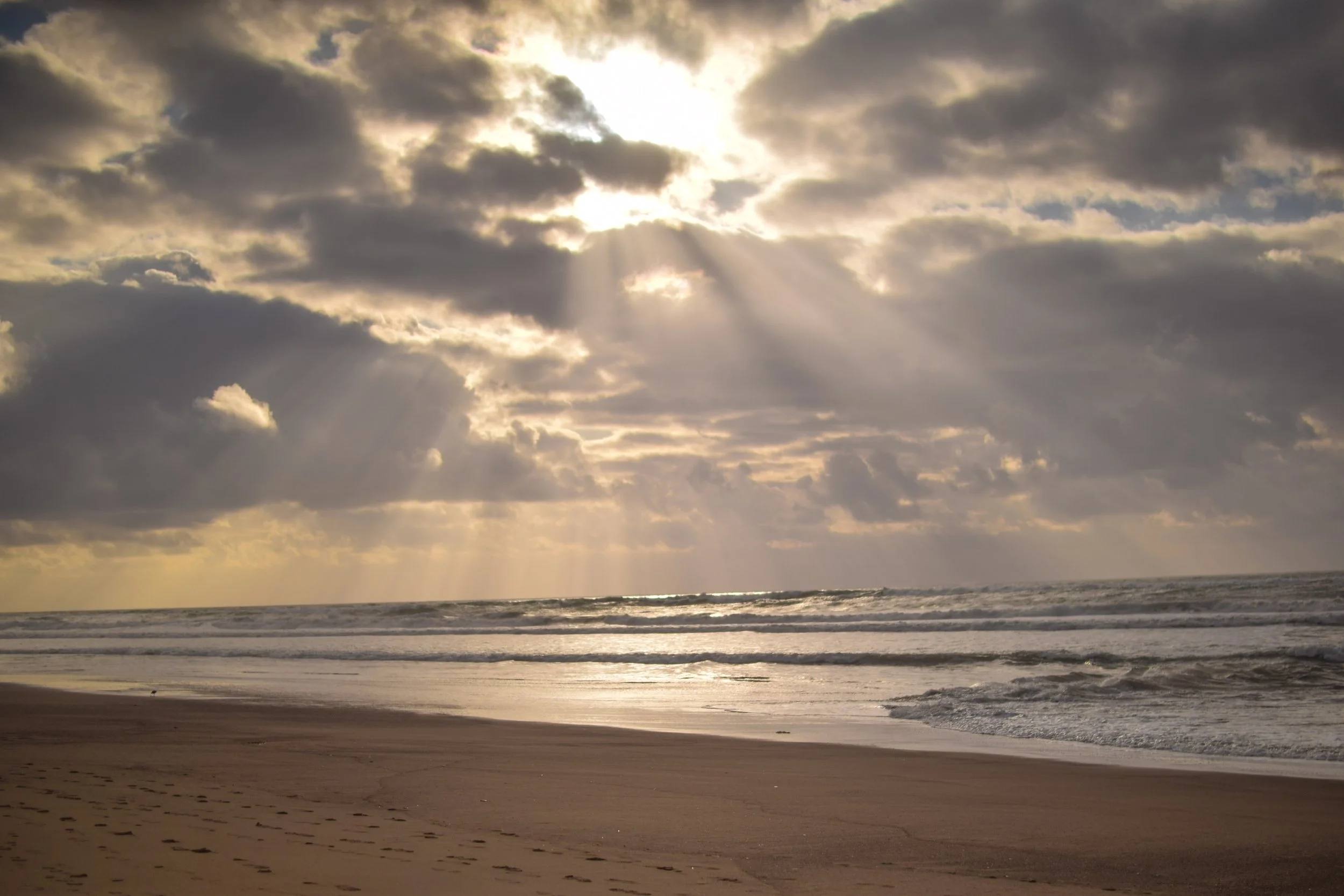 Sun rays breaking through cloudy sky over a calm beach with footprints in the sand.