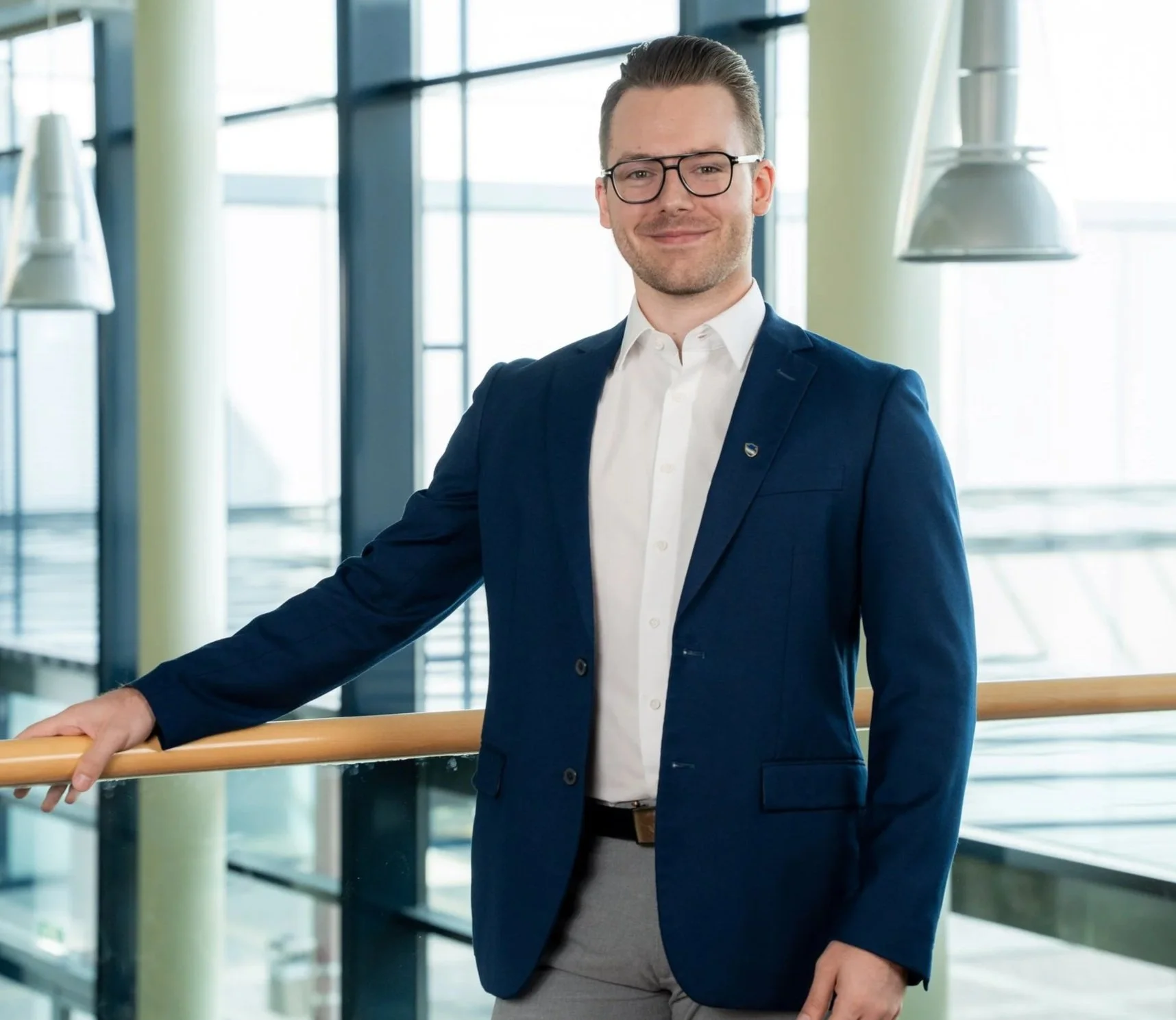 Ein Mann trägt eine Brille, dunkle Kleidung, eine weiße Hemd und einen blauen Blazer. Er steht in einem modernen Gebäude mit großen Fenstern und einem Handlauf, lächelt und blickt in die Kamera.