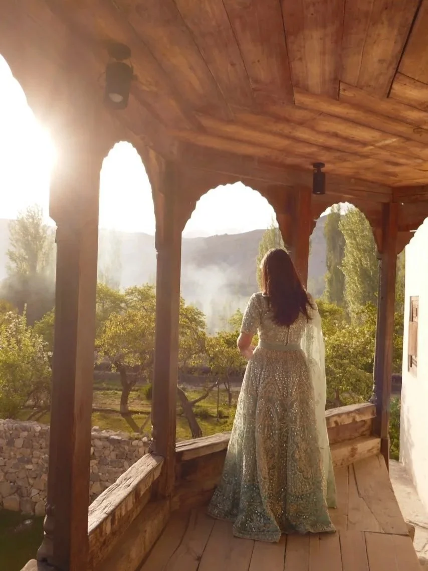 Woman in a wedding dress standing on a rustic wooden balcony overlooking a lush, green landscape with trees and mountains in the distance during sunset.