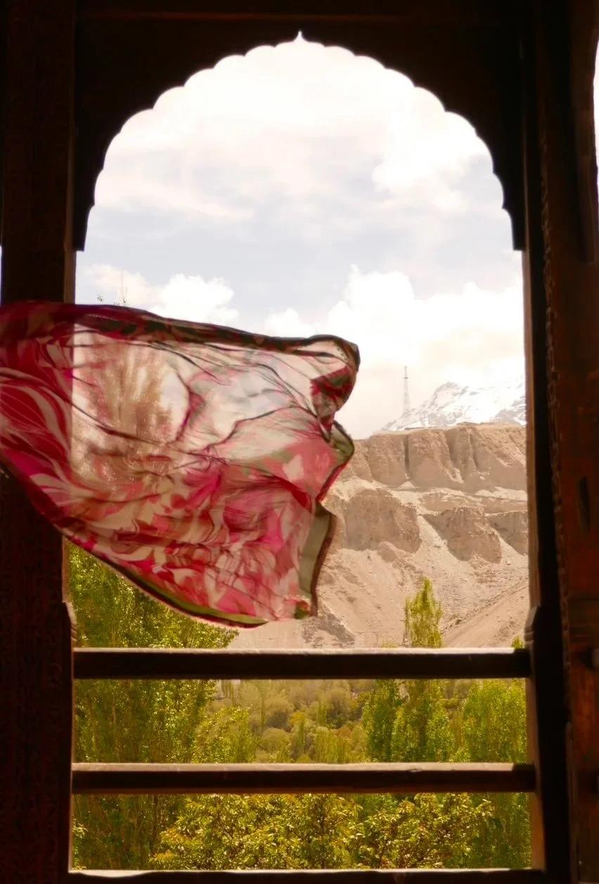 View through a window with wooden frame, showing a pink duppatta flowing in the wind, a landscape with green trees, brown rocky hills and snow-capped mountains, and a cloudy sky.