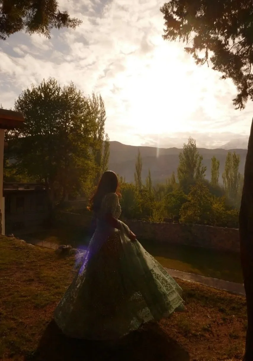 A woman in a wedding dress standing outdoors at sunset with mountains and trees in the background.