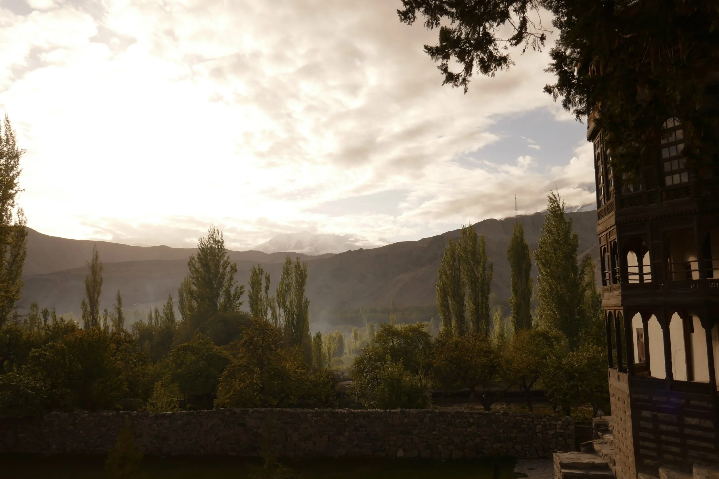 A scenic landscape with mountains in the background, trees in the foreground, and a partly cloudy sky during sunset, with an ornate building partially visible on the right side.