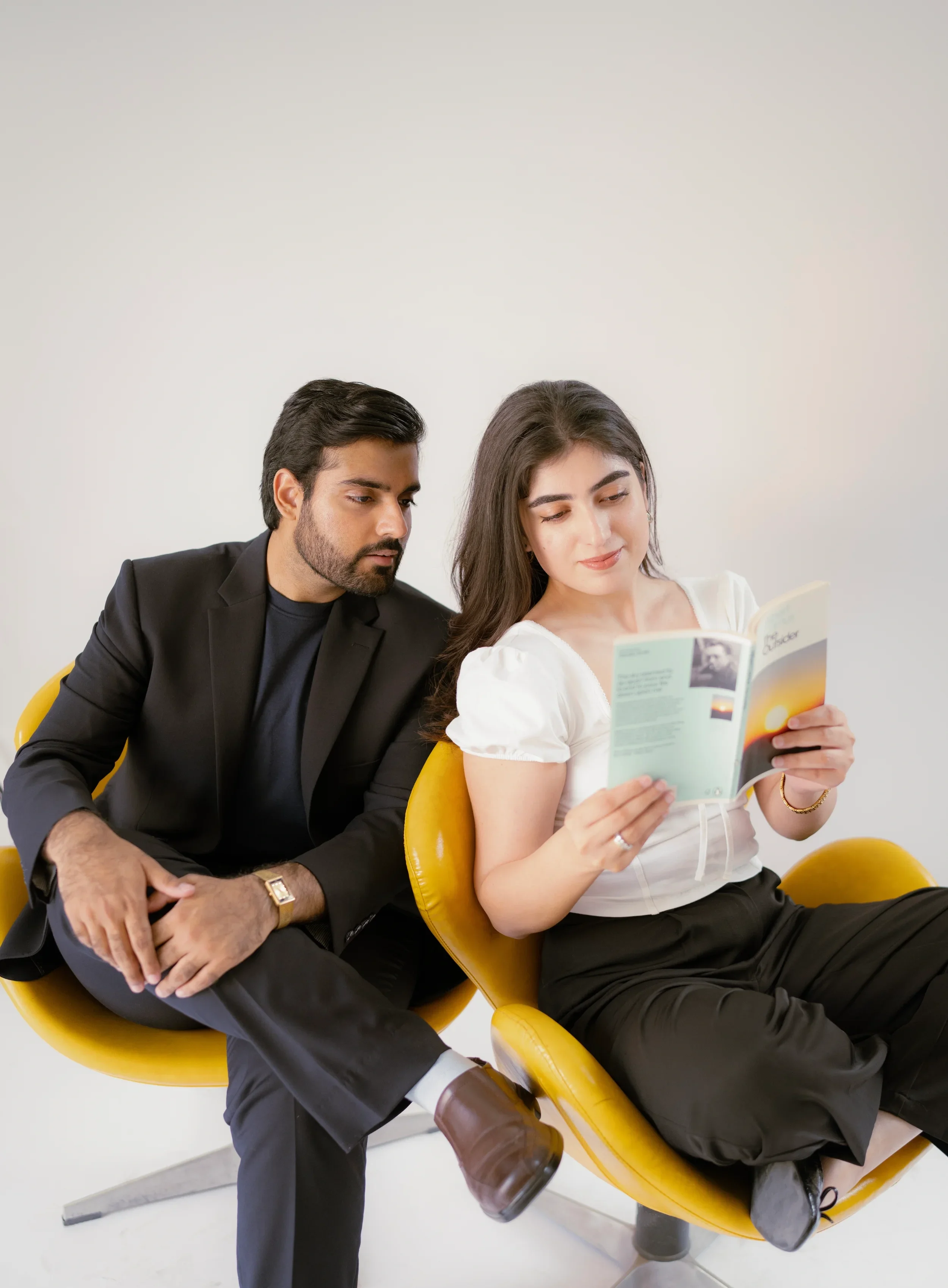 A man in a black suit and a woman in a white top and black pants sitting on yellow chairs, with the man leaning towards the woman who is reading a book, both against a plain white background.