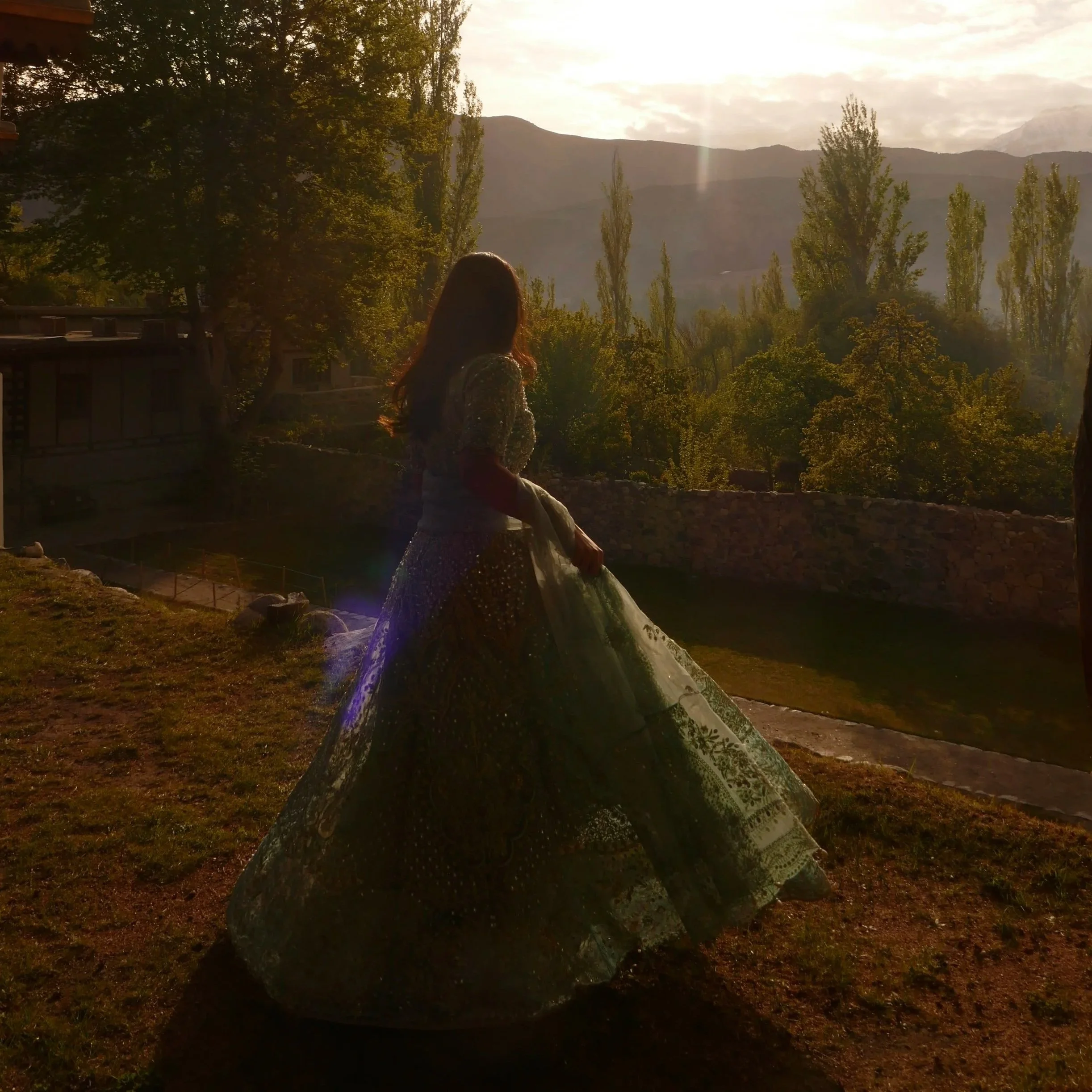 Person with long hair sitting outdoors on a stone bench, looking at a scenic mountain view with trees and cloudy sky during sunset.
