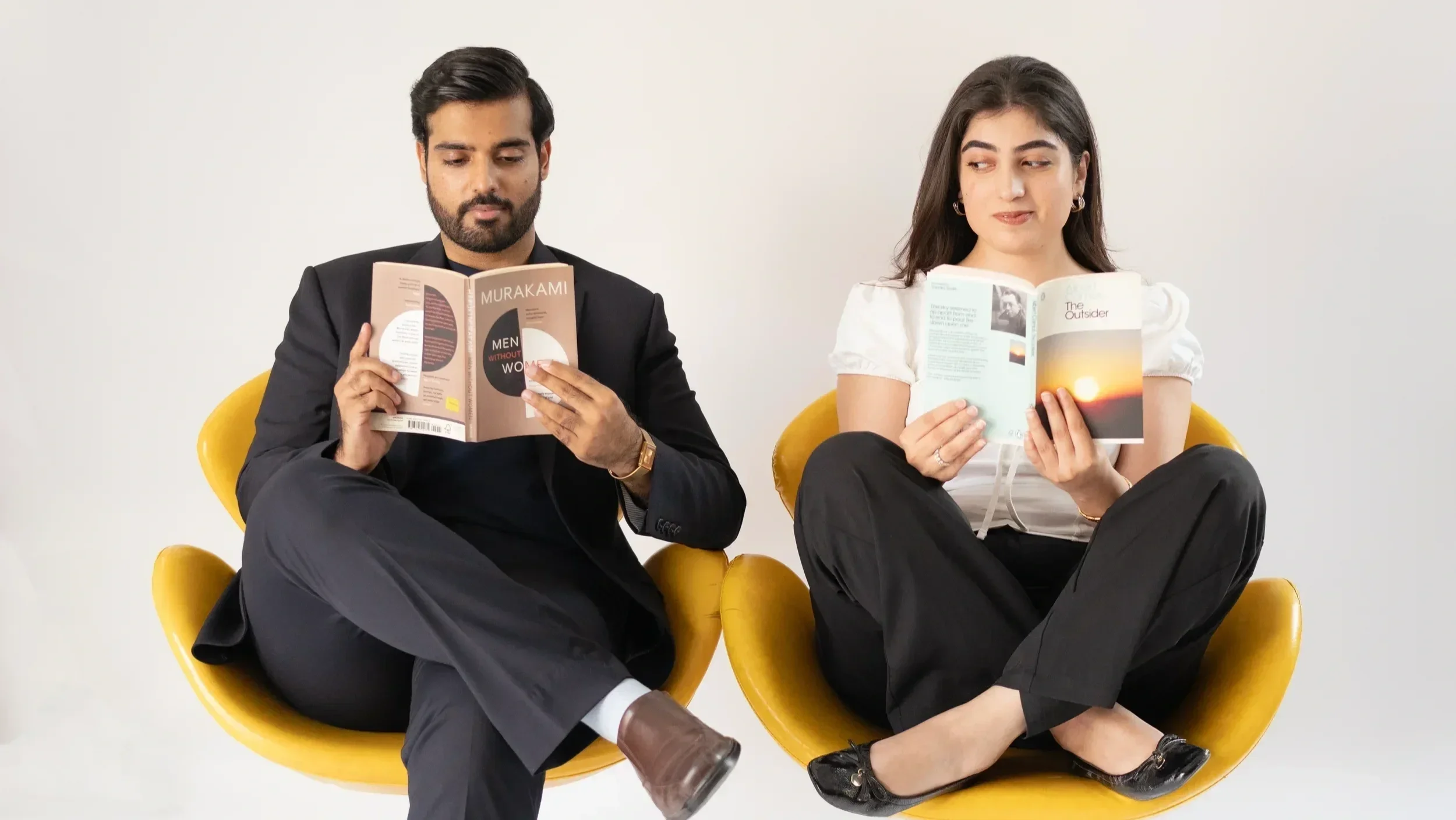 A man in a black suit and a woman in a white top sit in yellow chairs, reading books, against a plain white background.