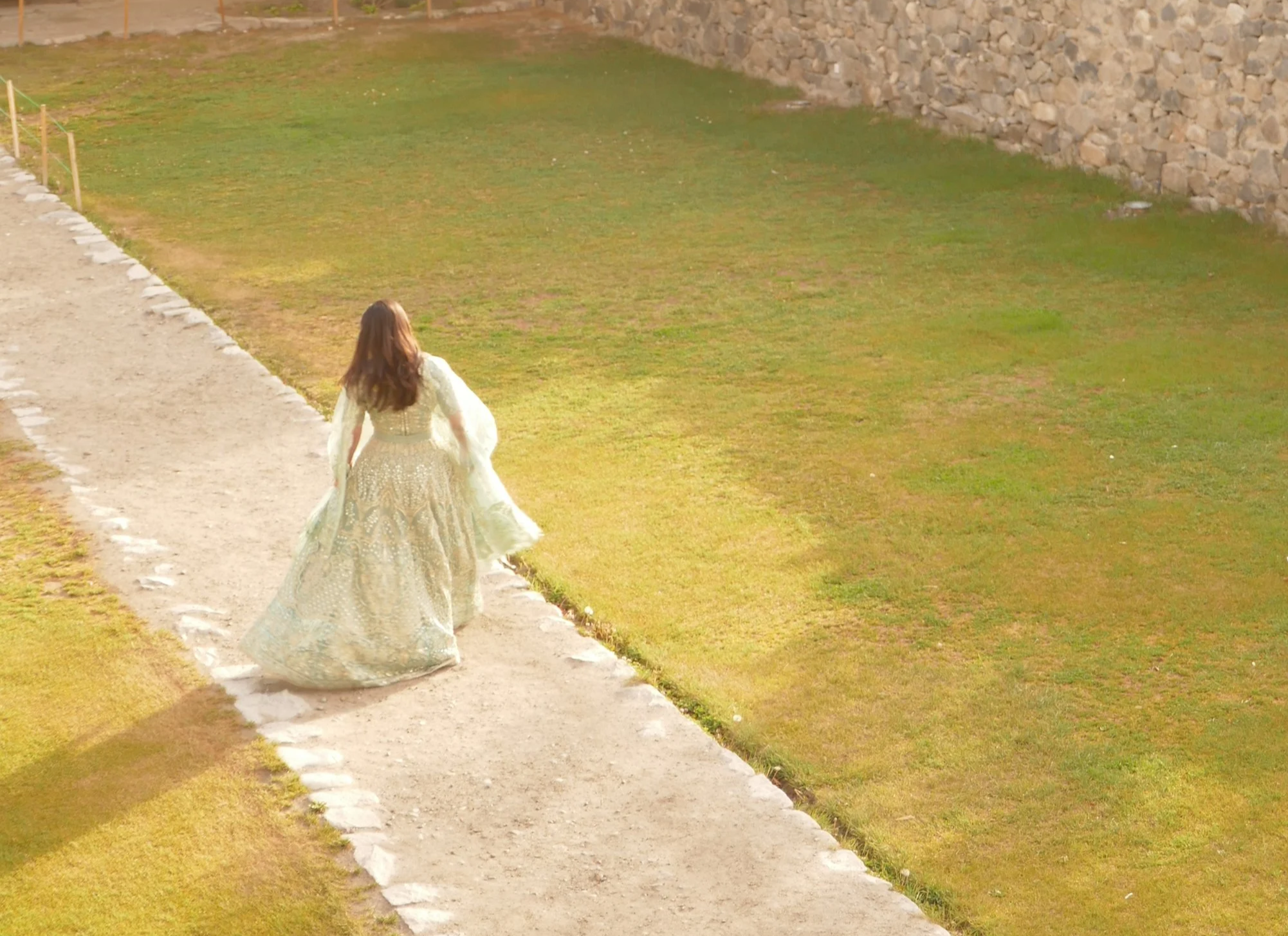 A woman in a flowing patterned gown walking along a gravel path beside a grassy area with a stone wall in the background.