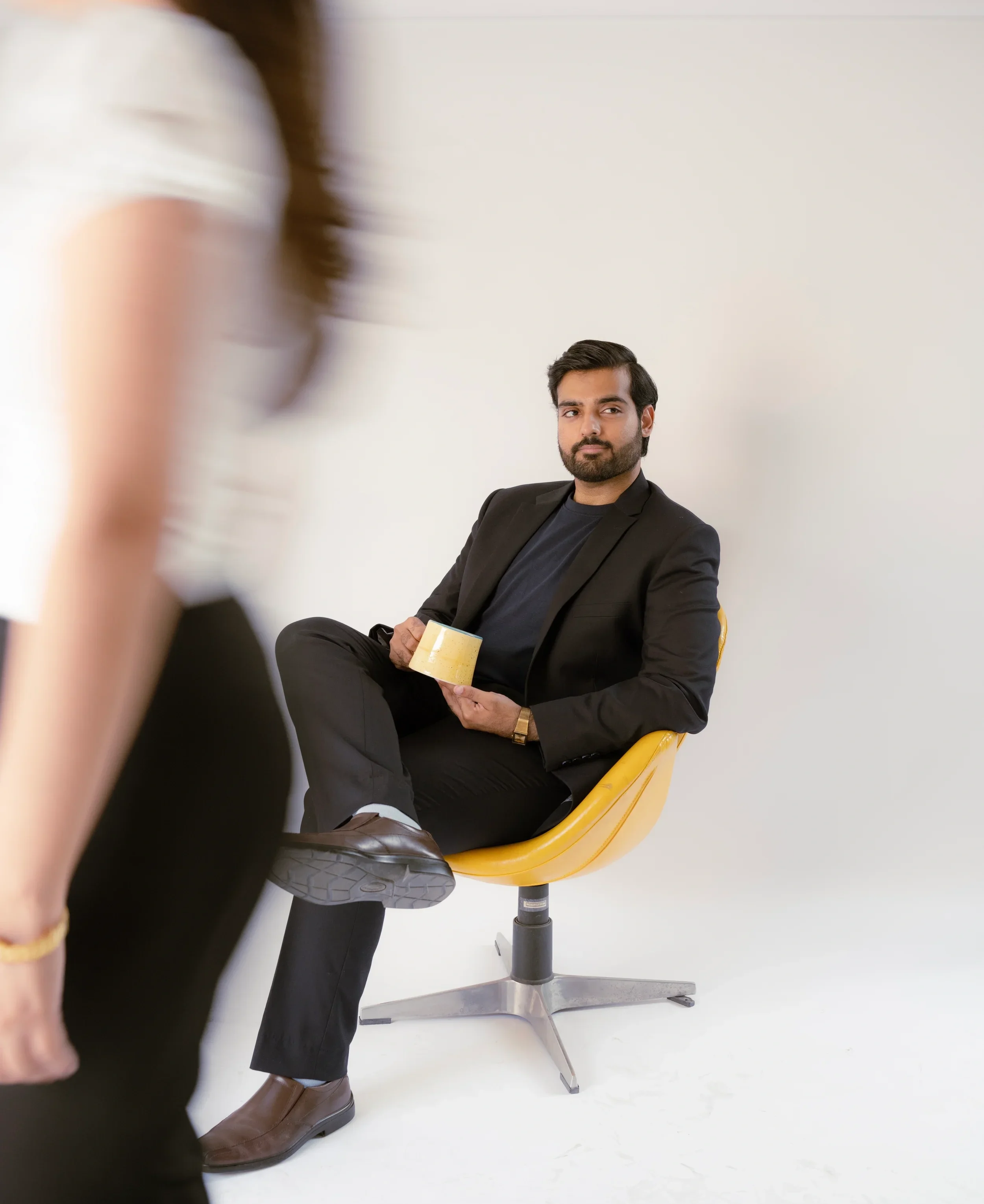 A man wearing a black blazer, dark shirt, and brown shoes sitting on a yellow chair, holding a beige coffee mug, with a white wall background.