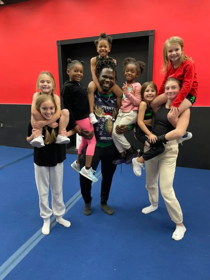 Group of children and a man at an indoor gymnastics or cheerleading gym, posing together with smiles. The background is red and black, and the floor is blue.