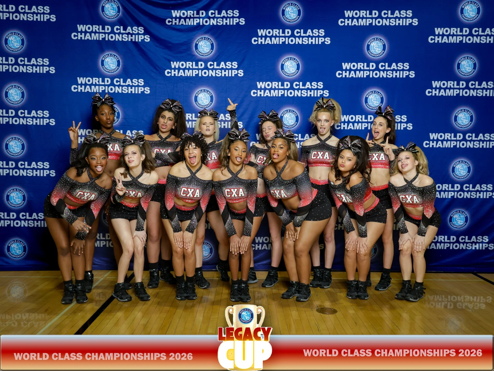 A team of cheerleaders in black and red uniforms with 'CXA' written on the front, posing together on a gym floor in front of a backdrop that says 'World Class Championships 2026' and features the World Cheerleading Alliance logo, with some cheerleaders making peace signs and others sticking out their tongues.