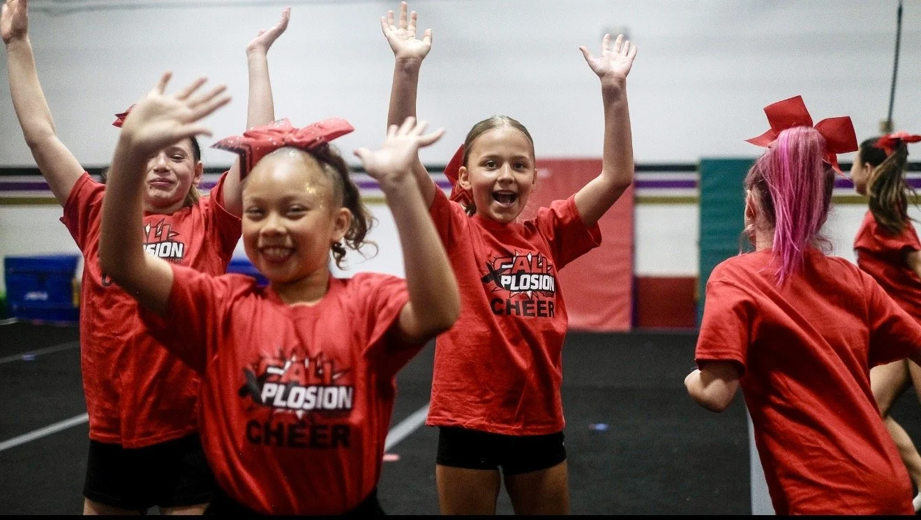 Cheerleaders celebrating during a cheer practice or performance, wearing red shirts with 'Cheer' written on them, inside a gym.