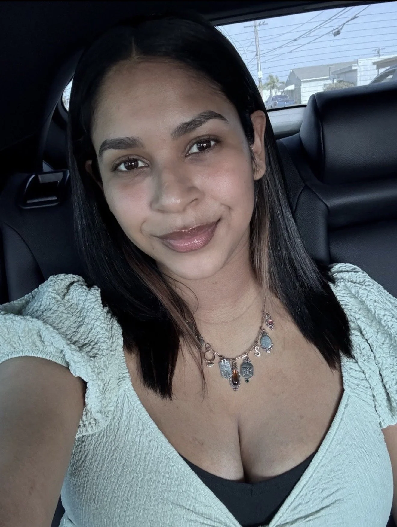 A woman with dark hair and makeup, wearing a necklace and a light-colored top, is sitting in the backseat of a car, smiling at the camera.