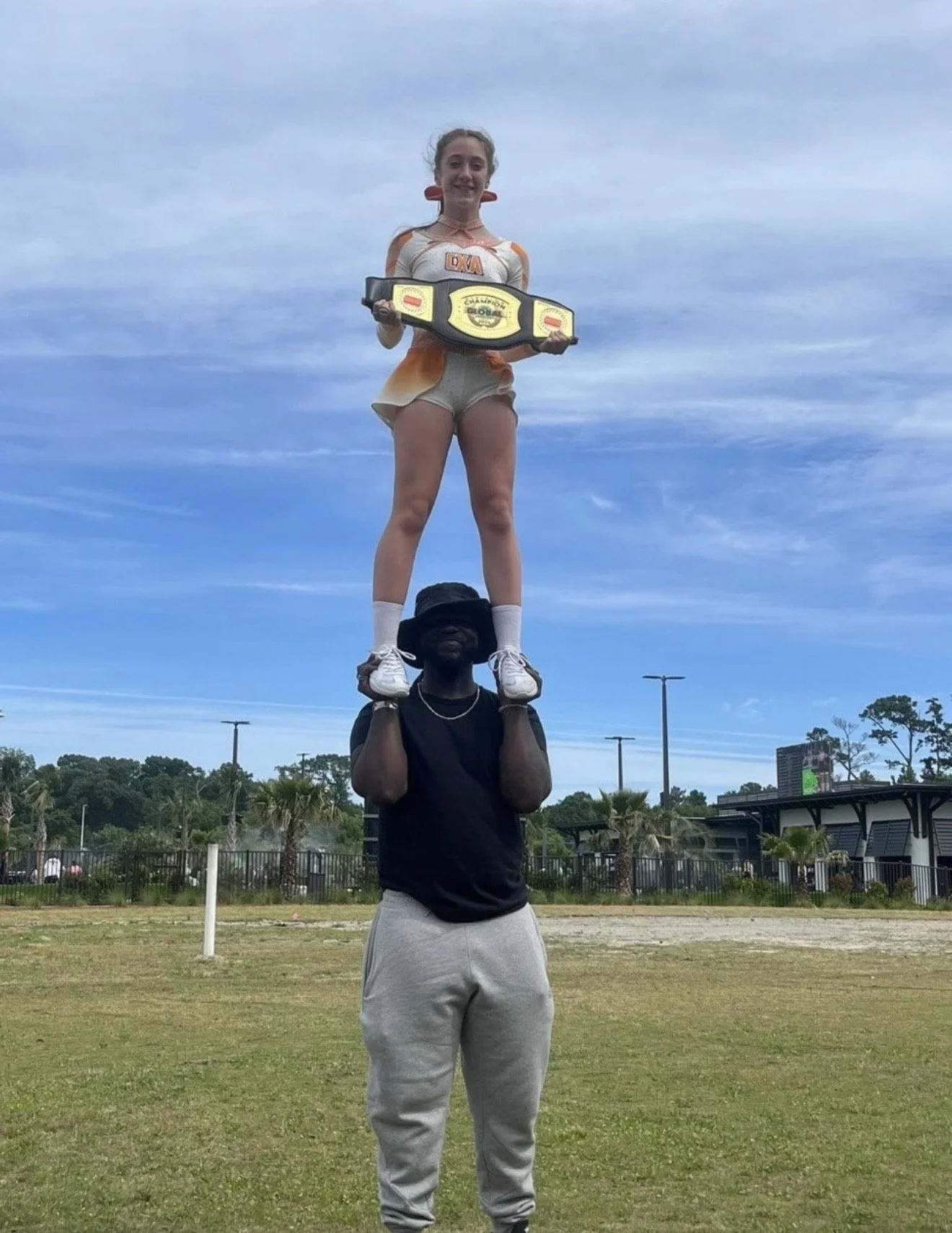 A woman standing on the shoulders of a man outdoors, holding a championship wrestling belt, with a sports stadium and trees in the background.