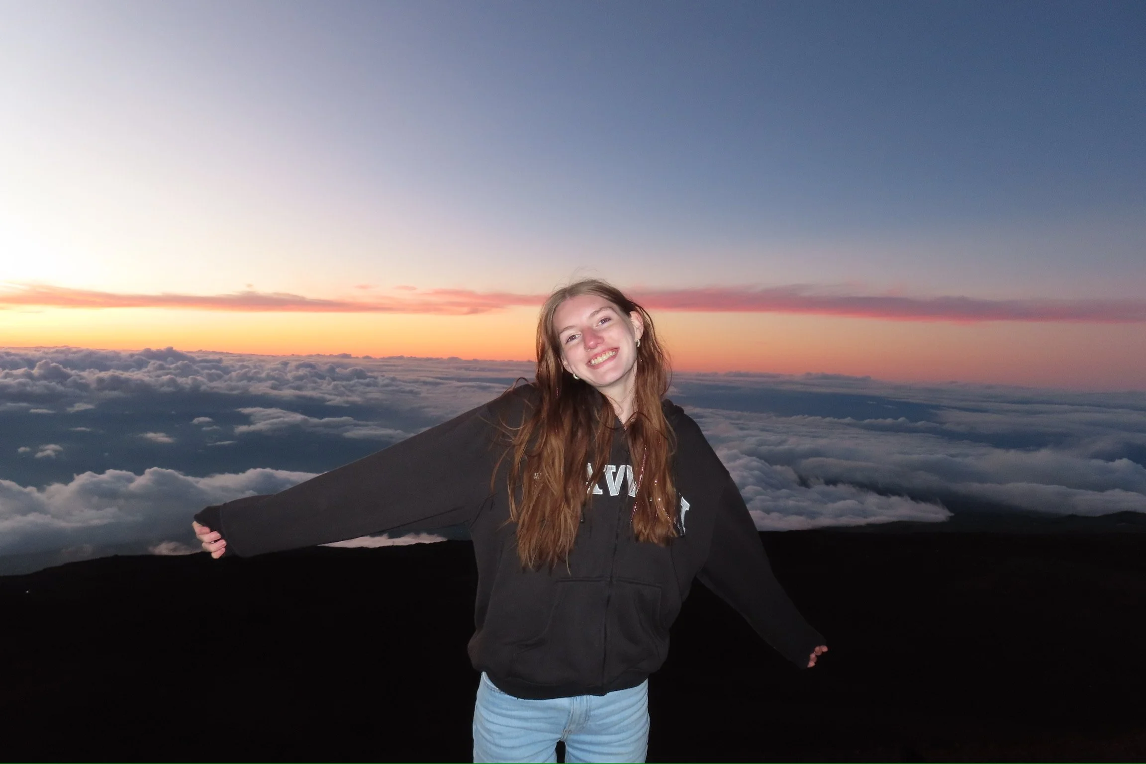 A young woman with long hair smiling and spreading her arms, standing outdoors at sunset or sunrise above the clouds.