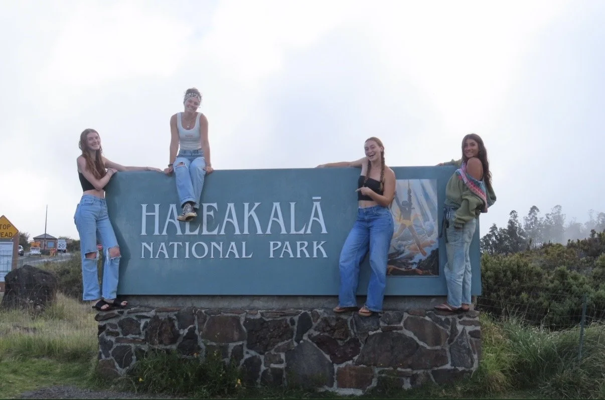 Four young women posing around the Haleakalā National Park sign, with some sitting and others standing, on a cloudy day.