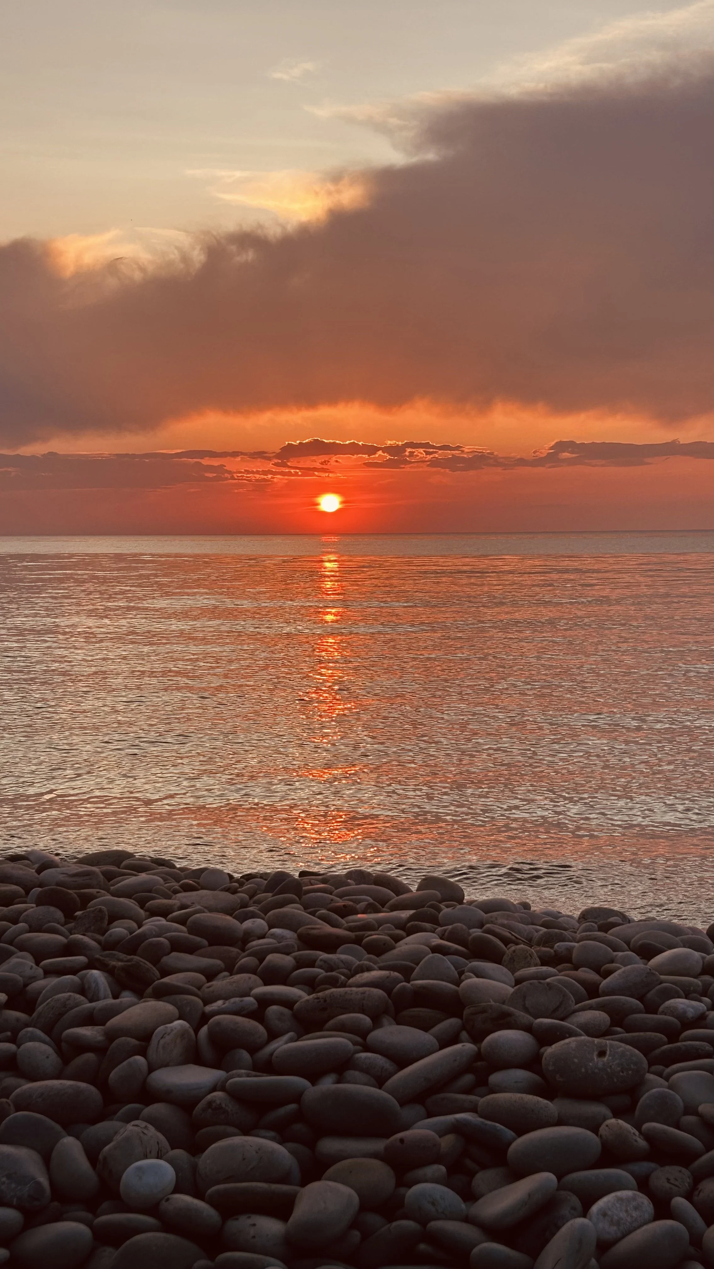Sunset over a calm ocean with a pebble beach in the foreground and colorful clouds in the sky.